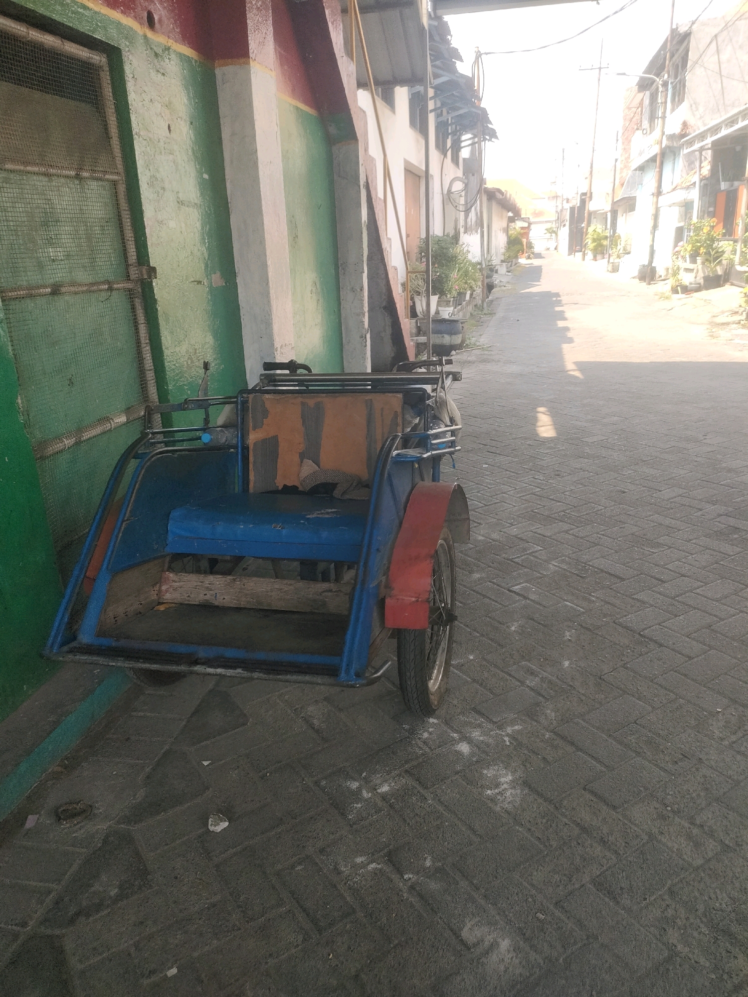 The image shows a blue and red trishaw parked on a brick-paved street next to a building with green and white walls. The building appears to be made of concrete and has a metal grate covering a window or opening. A narrow street lined with buildings and potted plants extends into the background, suggesting an urban environment in a warm climate. The lighting indicates it is daytime, with sunlight casting shadows on the street. The trishaw is unoccupied and appears to be in a residential or commercial area. Given the architecture and prevalence of trishaws, the location could be a street in Surabaya, Indonesia, a city known for its dense urban landscape and traditional modes of transport.
