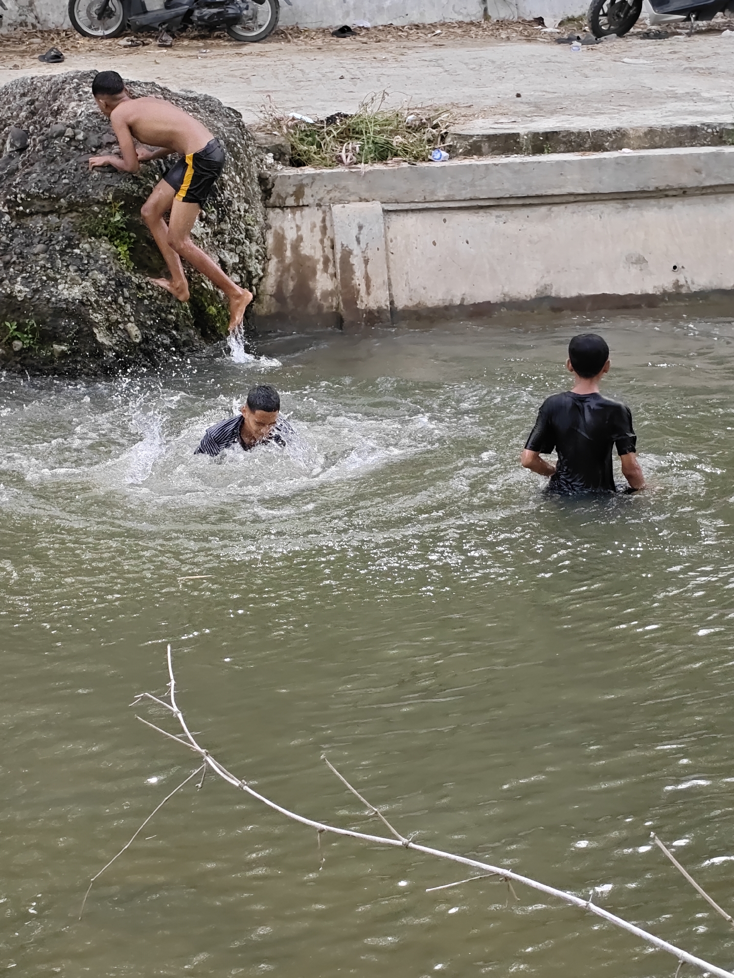 In Saweuk, Indonesia, three young males are engaged in recreational activity in a shallow, murky body of water adjacent to a rocky embankment and a concrete structure. On the left, a boy with dark hair, wearing black swim shorts with yellow accents, is in the process of jumping from the rocky embankment into the water, with his legs extended downward and his arms braced against the rocks. Water splashes upwards from his feet. In the center, another boy, also with dark hair, is swimming or playing in the water, creating significant splashes around him. He is wearing a dark, striped shirt. To the right, a third boy, facing away from the camera, stands in the water up to his waist. He has dark hair and is wearing a wet black t-shirt. The environment appears to be an urban or semi-urban setting, with a concrete walkway visible behind the embankment, where a motorcycle is parked. The water surface reflects sunlight, and a few dry branches are visible in the foreground, partially submerged in the water.