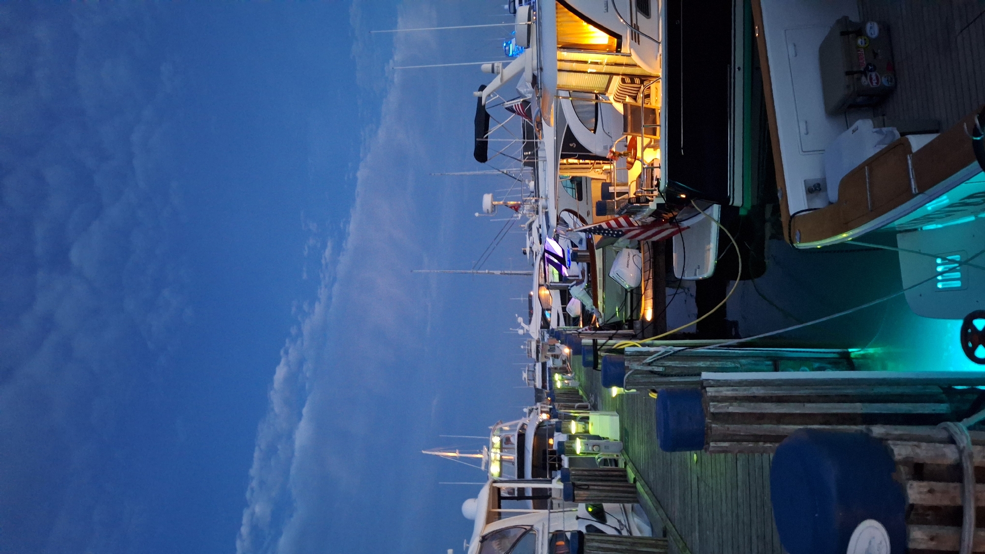 The image depicts a twilight scene at a marina in Nantucket, United States of America, showcasing numerous yachts docked along a wooden pier. The sky is a deep blue with wispy clouds, indicating the transition from day to night. The boats are illuminated by interior lights and underwater blue and green lights, casting a serene glow on the water. An American flag is prominently displayed on one of the yachts. The pier itself is made of weathered wood planks and is lined with bollards. The overall atmosphere is one of tranquility and calm as the boats rest in the harbor.