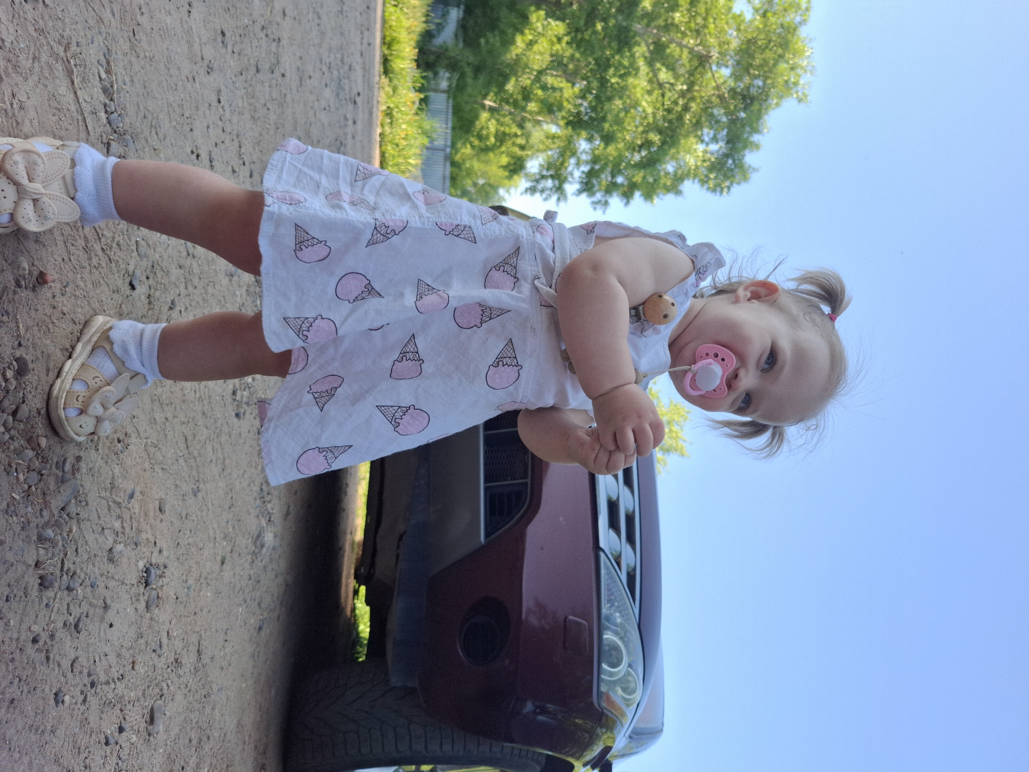 A toddler is standing next to a car on a dirt and gravel surface, with a partial view of a tree and fence in the background under a blue sky. The child is wearing a white dress patterned with pink ice cream cones, white socks, and light-colored sandals. They have a pink pacifier in their mouth. The car is maroon, with part of the front grill and headlights visible. The location cannot be determined based on this image alone.
