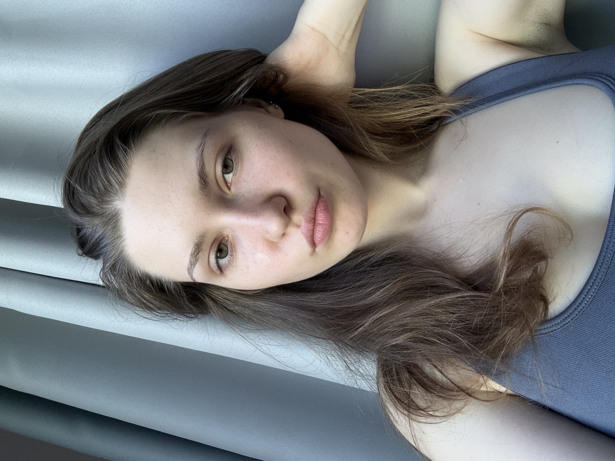 A young adult woman is shown in a close-up, high-angle shot, lying down and looking directly at the camera. The image is rotated, positioning her head towards the top of the frame. She has fair skin with some freckles, light-colored eyes (appearing hazel or greenish-brown), and natural light pink lips. Her expression is calm and neutral. Her medium-brown hair, with lighter highlights, falls loosely around her face and shoulders. A small silver-colored stud piercing is visible on the cartilage of her left ear. She is wearing a dark gray or slate blue athletic-style top or bra with a visible strap and ribbed detailing. Her right arm is raised and bent above her head. The background is composed of soft, light gray or blue-gray fabric, possibly curtains or folded material, creating gentle folds and textures. The scene is illuminated by bright, natural light, suggesting it is daytime. No text is visible in the media file. The location context is Saint Petersburg, Russia.