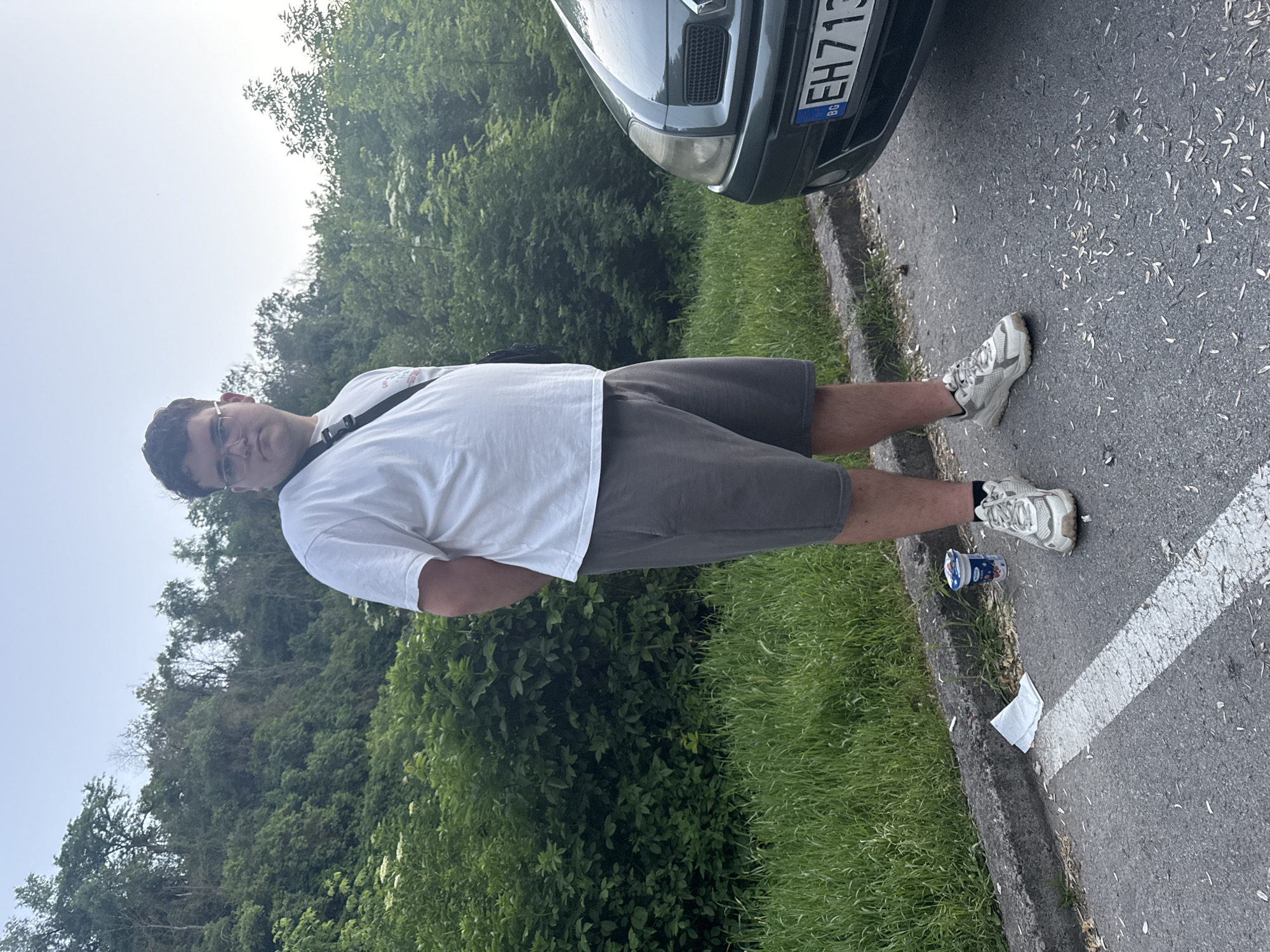 The photograph shows a man standing on a paved surface next to a car, with foliage and trees in the background. The man is wearing glasses, a white t-shirt, gray shorts, and white sneakers. The car is a dark color, and the license plate reads "EH 713 BG". Litter, including a small yogurt container and a white tissue, is visible near the edge of the pavement. The location may be in Bulgaria, as suggested by the license plate country code. The photo does not provide definitive indicators to confirm the specific city of Pleven, or the presence of landmarks that are unique to that city.