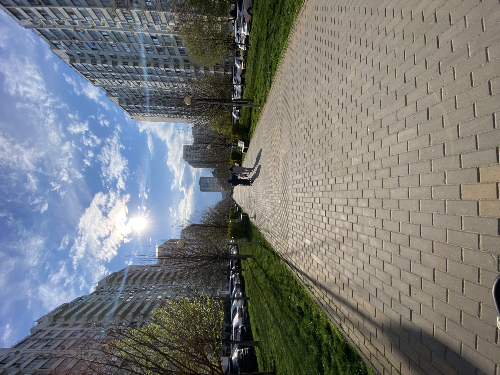 The image shows a paved pathway in an urban setting. Tall apartment buildings line either side of the pathway. A strip of green grass separates the pathway from a row of parked cars on each side. Several people are walking along the path in the distance. The sky is visible between the buildings and is blue with scattered clouds. The sun is shining brightly. The city is Unknown, Unknown.