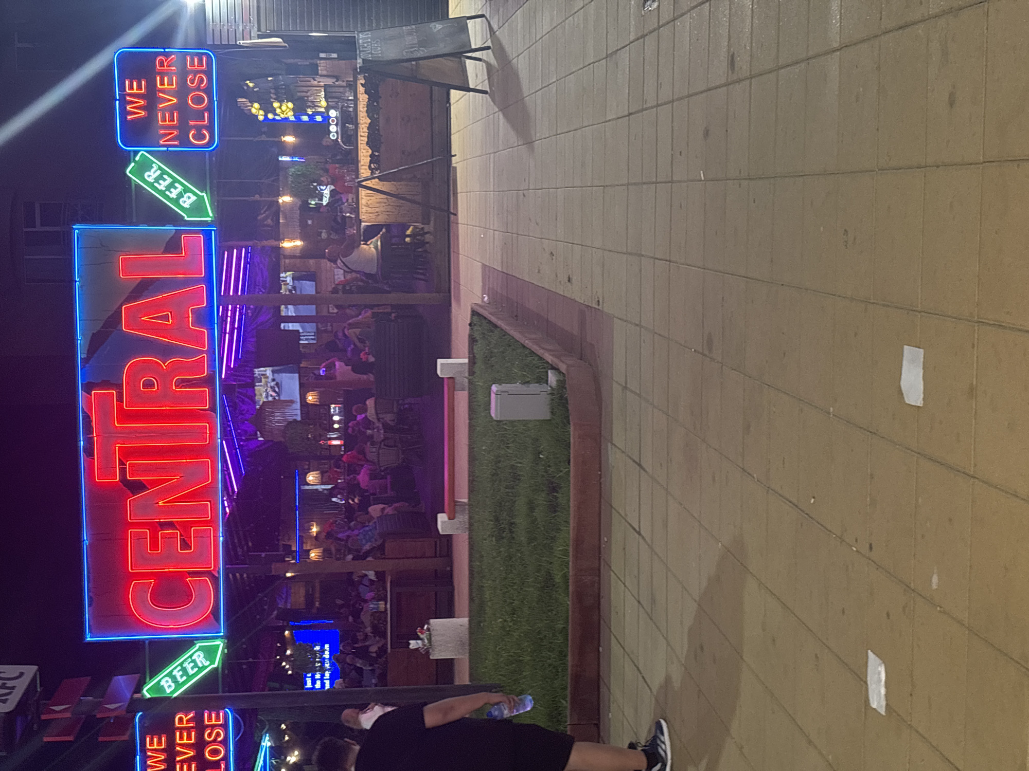 The image captures a nighttime street scene in Nesebar, Bulgaria, likely near a nightlife establishment. A prominent neon sign reading "CENTRAL" in red letters with a blue outline dominates the upper center of the frame. To its left, another neon sign in green with a blue outline says "BEER" with an arrow pointing downwards, and further left, a sign reads "WE NEVER CLOSE" in red neon. The background reveals a lively outdoor seating area with numerous patrons seated at tables and a bar. Several people are visible, some standing and others seated, engaged in various activities like drinking and socializing. The environment appears to be a pedestrian walkway paved with square tiles, with a strip of grass and a concrete planter separating the walkway from the establishment. Lights from the bar and surrounding area illuminate the scene.