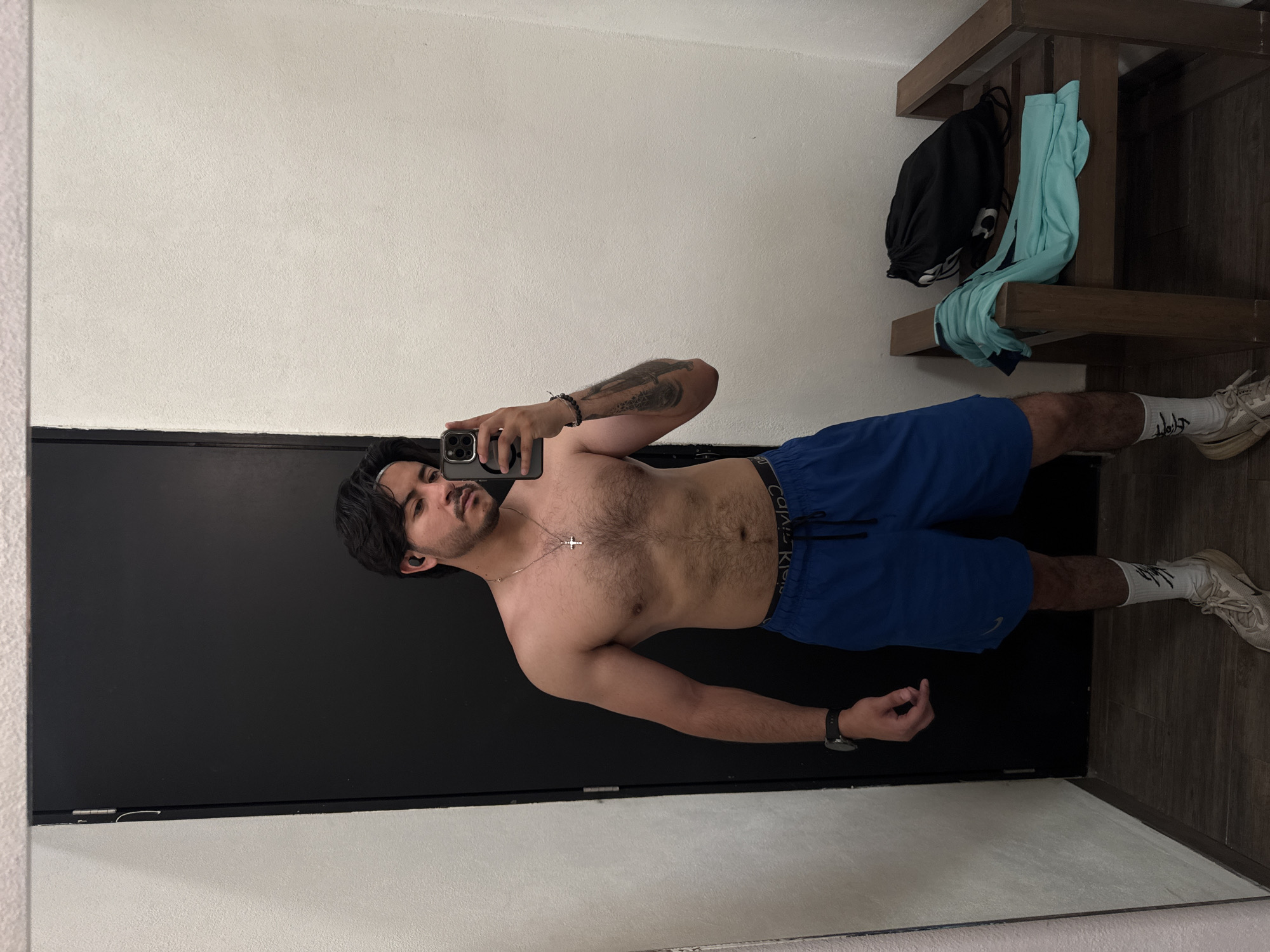 A shirtless man wearing blue athletic shorts and white sneakers is standing in what appears to be a fitting room, taking a selfie in a mirror. He has a tattoo on his left arm, a cross necklace, and is wearing a watch on his left wrist. A black bag and a light blue shirt are on a wooden shelf behind him. There is a door beside him. There is no indication of the city of Puebla, Mexico.