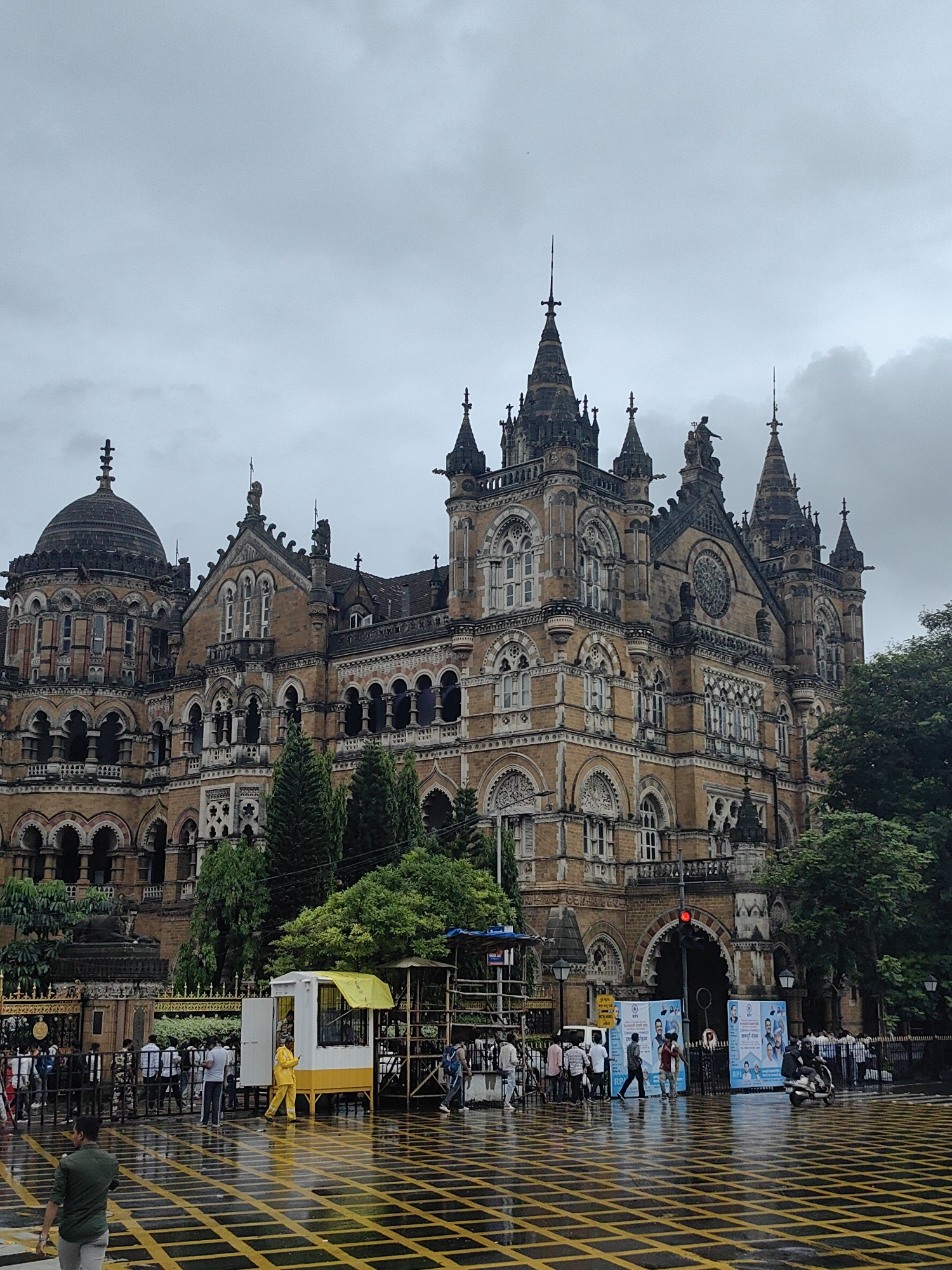 The image depicts the Chhatrapati Shivaji Maharaj Terminus (CSMT) in Mumbai, India, a historic railway station and a UNESCO World Heritage Site. The grand Victorian Gothic Revival architecture of the building is prominent, with its intricate stonework, domes, spires, and arched windows. It is a cloudy day, and the ground appears wet, suggesting recent rain, with yellow and black lines marked on the pavement.
In the foreground, several people are visible, some walking and others standing. A yellow and white booth with a yellow awning is present on the left side of the image, with a person in a yellow uniform standing outside. Further back, a crowd is gathered near the entrance of the station. Several vehicles, including a motorcycle and a few cars, are also present on the road. Lush green trees frame the right side of the building. The overall scene conveys a busy urban environment with a strong historical architectural presence, possibly during a period of public activity or transit.