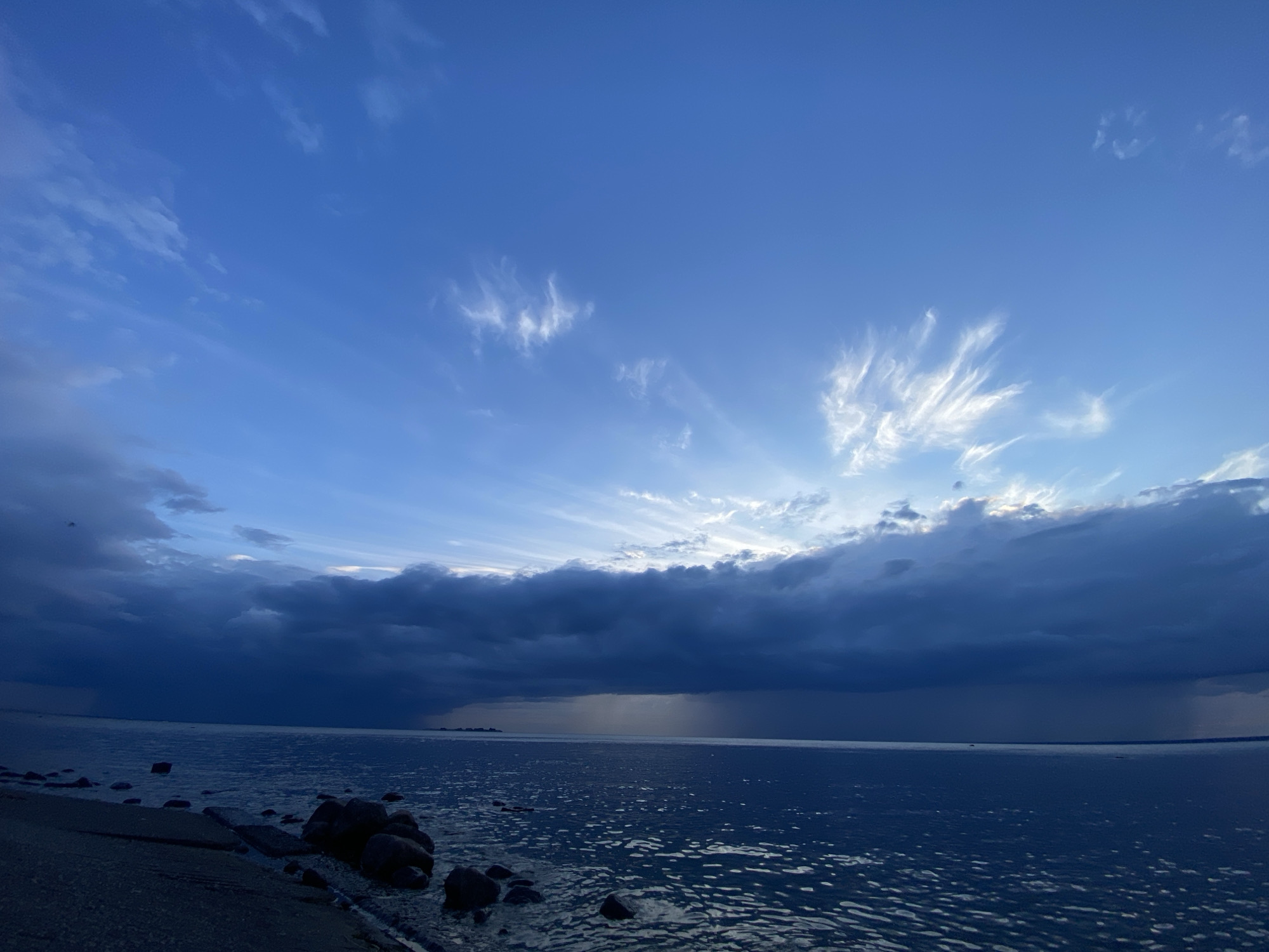 This image captures a stormy seascape in Sestroretsk, Russia. The sky is a dramatic display of deep blues and dark clouds, with streaks of sunlight breaking through in the upper right quadrant, creating visible rays. Wispy white clouds are scattered across the brighter parts of the sky. Below, a calm, dark body of water stretches to the horizon, reflecting the moody sky. The foreground features a rocky shoreline, with dark, wet stones and pebbles lining the edge of the water. A patch of dark, possibly wet sand, is visible to the left. A faint rain shower is depicted as a hazy curtain falling over the distant water. The scene conveys a sense of impending weather change, with the storm clouds gathering over the sea.