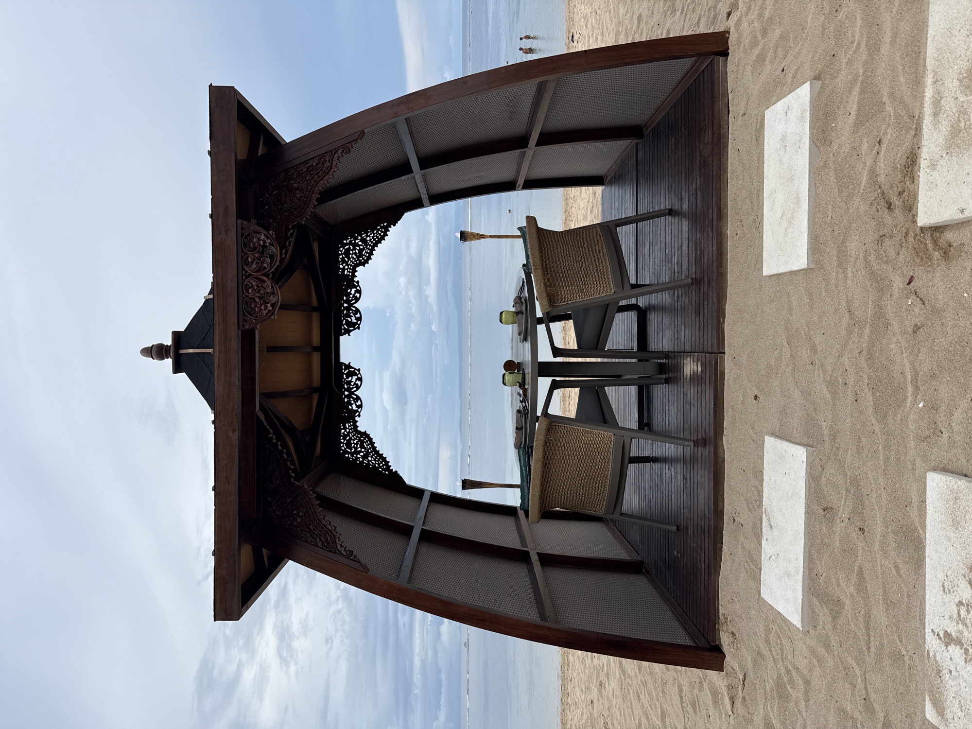 The image shows a dining setup in an open-air structure, likely a cabana or gazebo, on a beach in Nusa Dua, Indonesia. The structure is made of dark wood with ornate carvings. Inside, there is a table set for a meal with two chairs. In the background, the ocean is visible with people wading in the water. The beach sand is light brown. White rectangular stone-like objects are nearby the structure on the sand. The sky is partially cloudy.