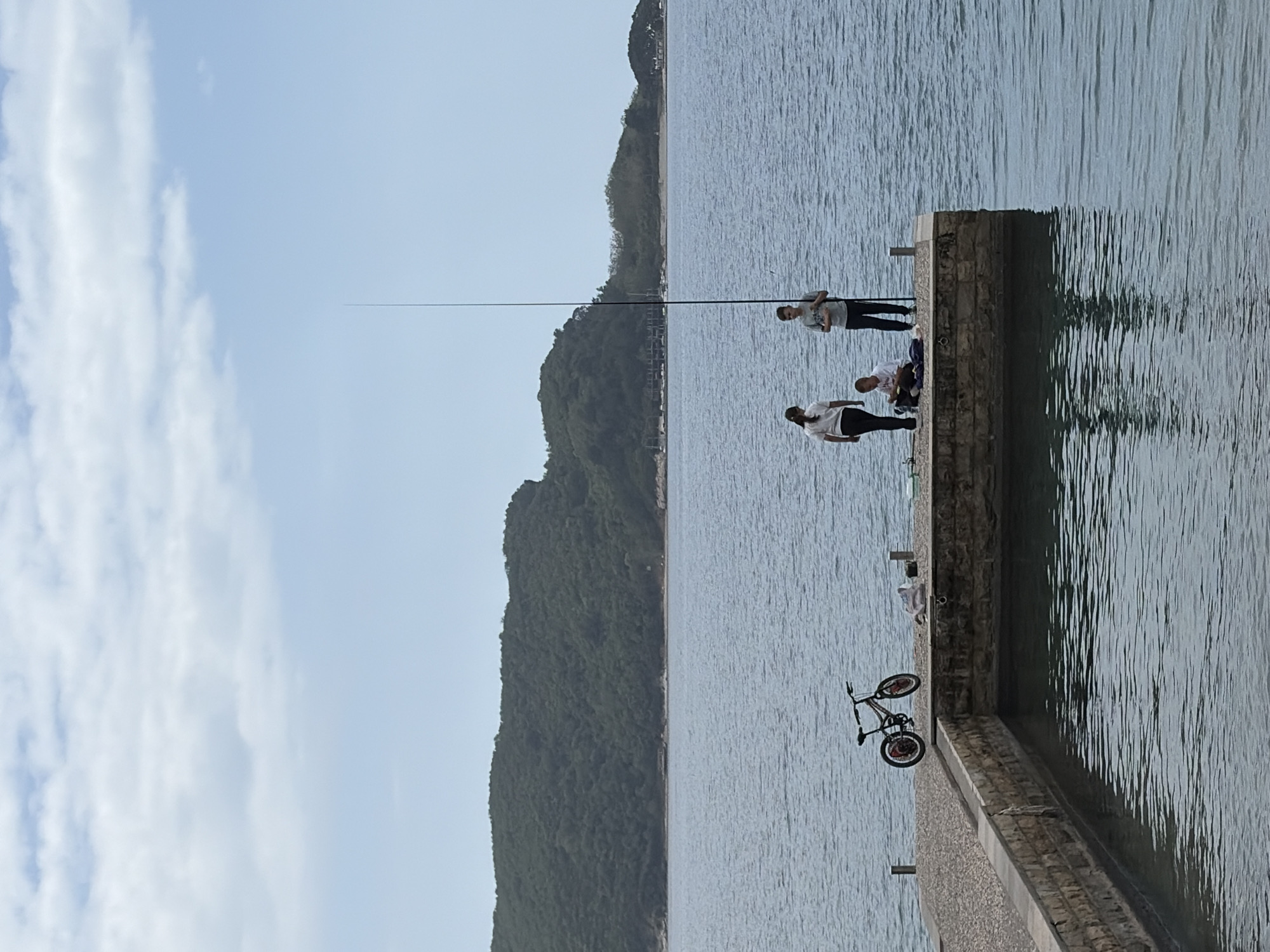 The scene is a sunny day in Budva, Montenegro. The location is a pier that extends into the water. Three people are on the pier, two standing and one sitting. A fishing rod is visible, extending out over the water. A bicycle rests against the pier. In the background is a tree-covered mountain. The water is calm. The sky is blue with some clouds.