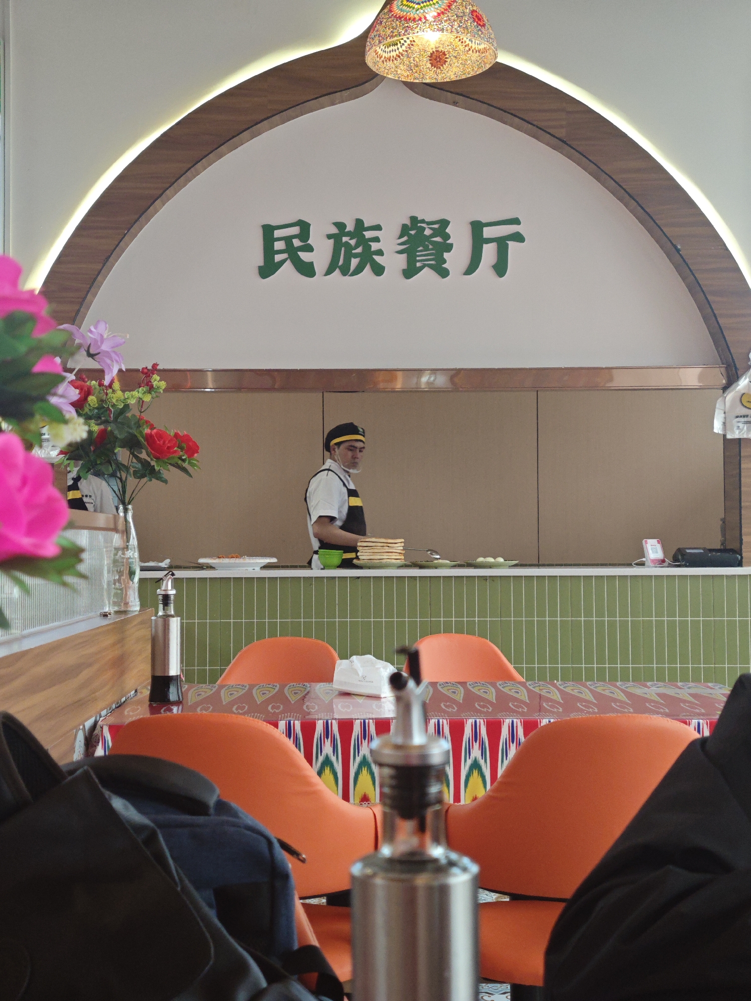 This image depicts the interior of a restaurant, identified by the Chinese characters "民族餐厅" (Mínzú Cāntīng), meaning "Ethnic Restaurant," prominently displayed in green above a serving archway. The scene appears to be set in Chongqing High-tech Zone, China.
In the background, a staff member, likely a chef or food preparer, stands behind a counter. This individual wears a white shirt, a black and yellow apron, and a black cap, with a face mask visible. On the counter, there's a stack of what appears to be flatbread or pancakes, a green bowl, and small plates holding food items, possibly dumplings or eggs. A card reader and another electronic device are also visible on the counter, which features a facade of green tiles.
The foreground showcases the dining area, featuring bright orange chairs with patterned fabric on their seatbacks, arranged around a table. The table is covered with a vibrant tablecloth displaying geometric and traditional patterns in red, yellow, green, and blue hues, suggesting an ethnic or regional cultural influence. On the table, two metallic dispensers, likely for oil and vinegar, and a packet of napkins are visible. A black backpack occupies the lower left corner, slightly out of focus. Artificial flowers, including pink, red, and purple varieties, are arranged in a clear glass vase on a nearby surface.
Above the counter, a decorative wooden archway with embedded lighting creates a warm glow. A colorful, mosaic-patterned lamp hangs from the ceiling in the upper right corner, contributing to the restaurant's distinct atmosphere. The lighting suggests it is daytime.