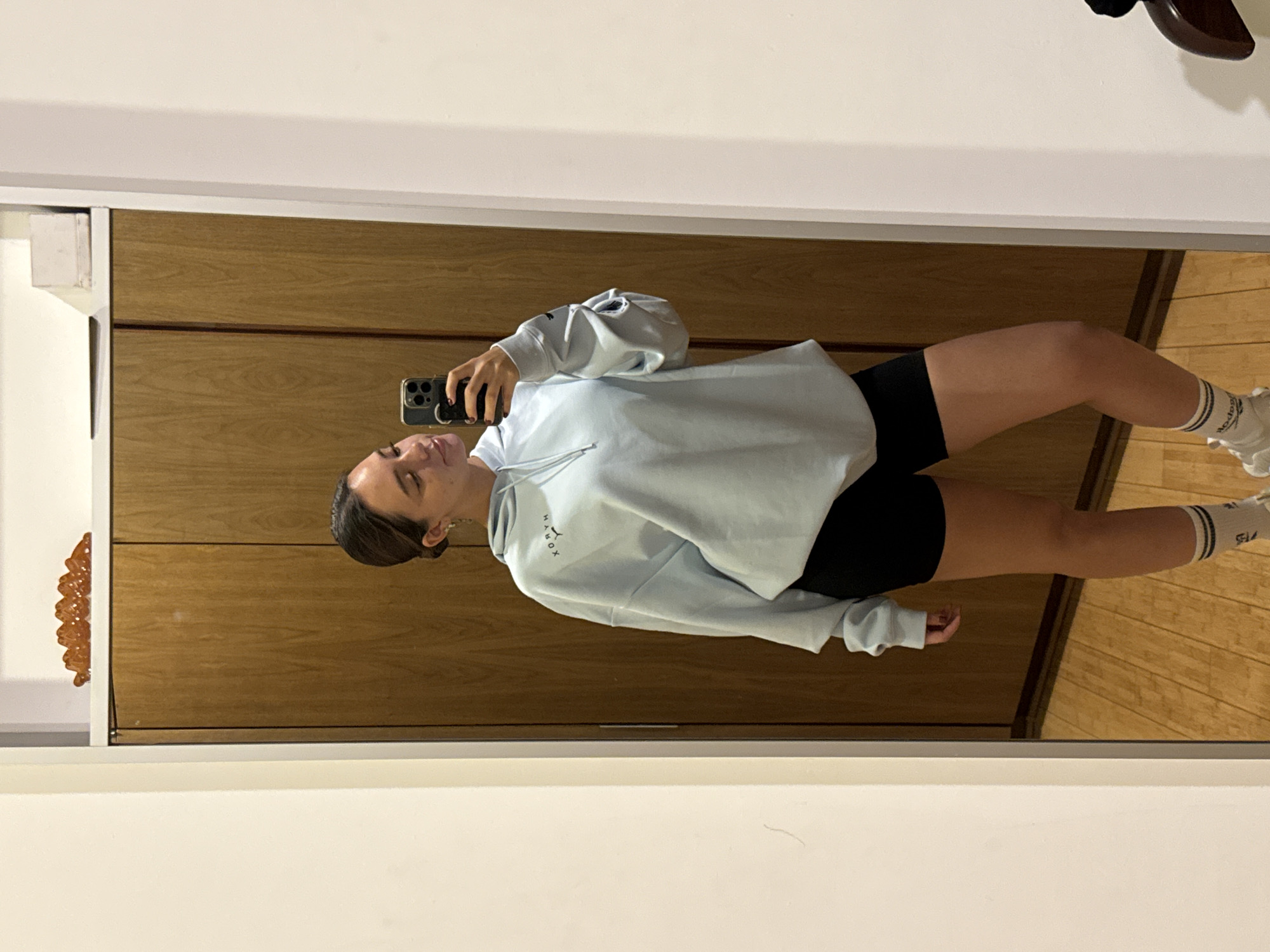 A woman is standing in front of a full-length mirror, taking a selfie with her phone. She is wearing a light blue hoodie and black shorts, with white socks and sneakers. The mirror reflects a wooden closet or cabinet behind her and a wooden floor. To the side of the closet is some sort of decorative object on the wall. The location is most likely inside a residence. There is no evidence indicating this photograph was taken in San Bernardino Tlaxcalancingo, Mexico.