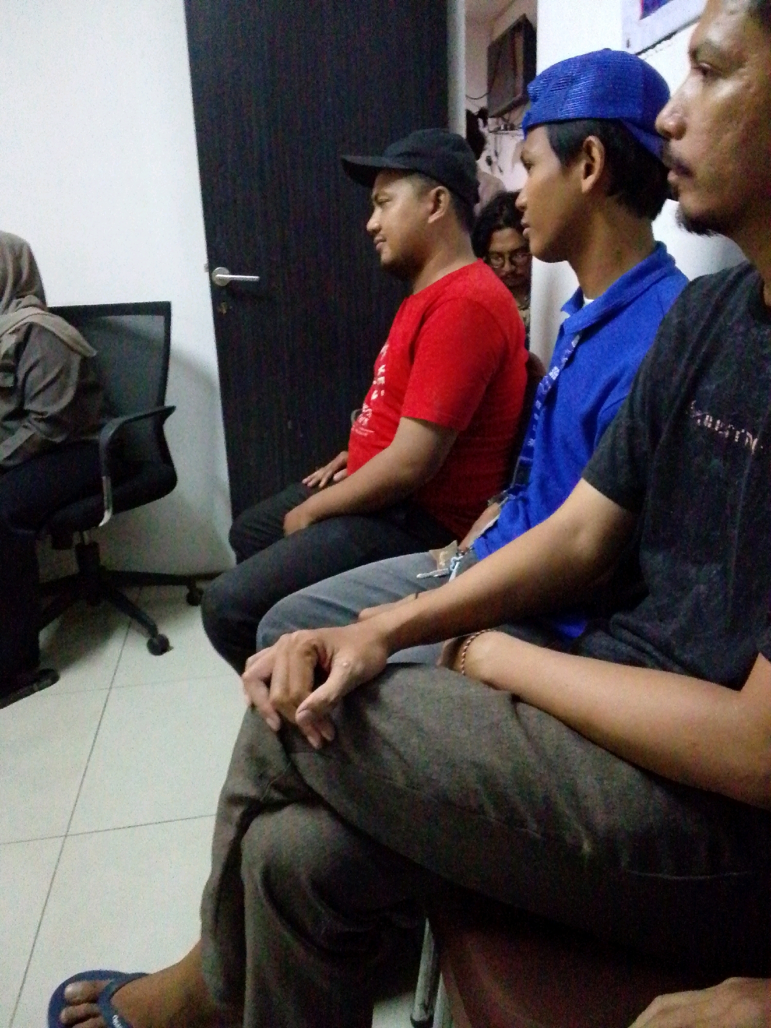 The image shows four men sitting in an indoor setting. From left to right, the first man is wearing a grey headscarf and grey clothing, and is partially visible. The second man is wearing a red t-shirt and a black baseball cap, and is looking to his left. The third man is wearing a blue t-shirt and a blue baseball cap, and is looking to his left. The fourth man is wearing a black t-shirt and is looking to his left. They appear to be sitting in chairs, with one chair visible on the left side of the frame. The background includes a white wall, a dark wooden door with a silver handle, and other indistinct objects. The flooring appears to be tiled. The context suggests a casual gathering or meeting. No specific location within North Jakarta, Indonesia, can be identified from the visual information.