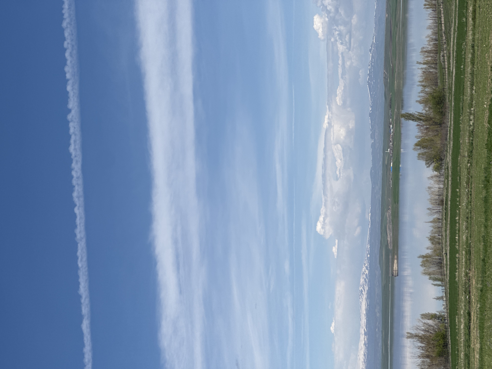 The image depicts a landscape in Aghin, Armenia, showcasing a body of water reflecting a mountainous terrain with snow-capped peaks. A line of trees borders the water's edge, separating it from a green grassy field. Above, the sky is blue with contrails and wispy clouds.