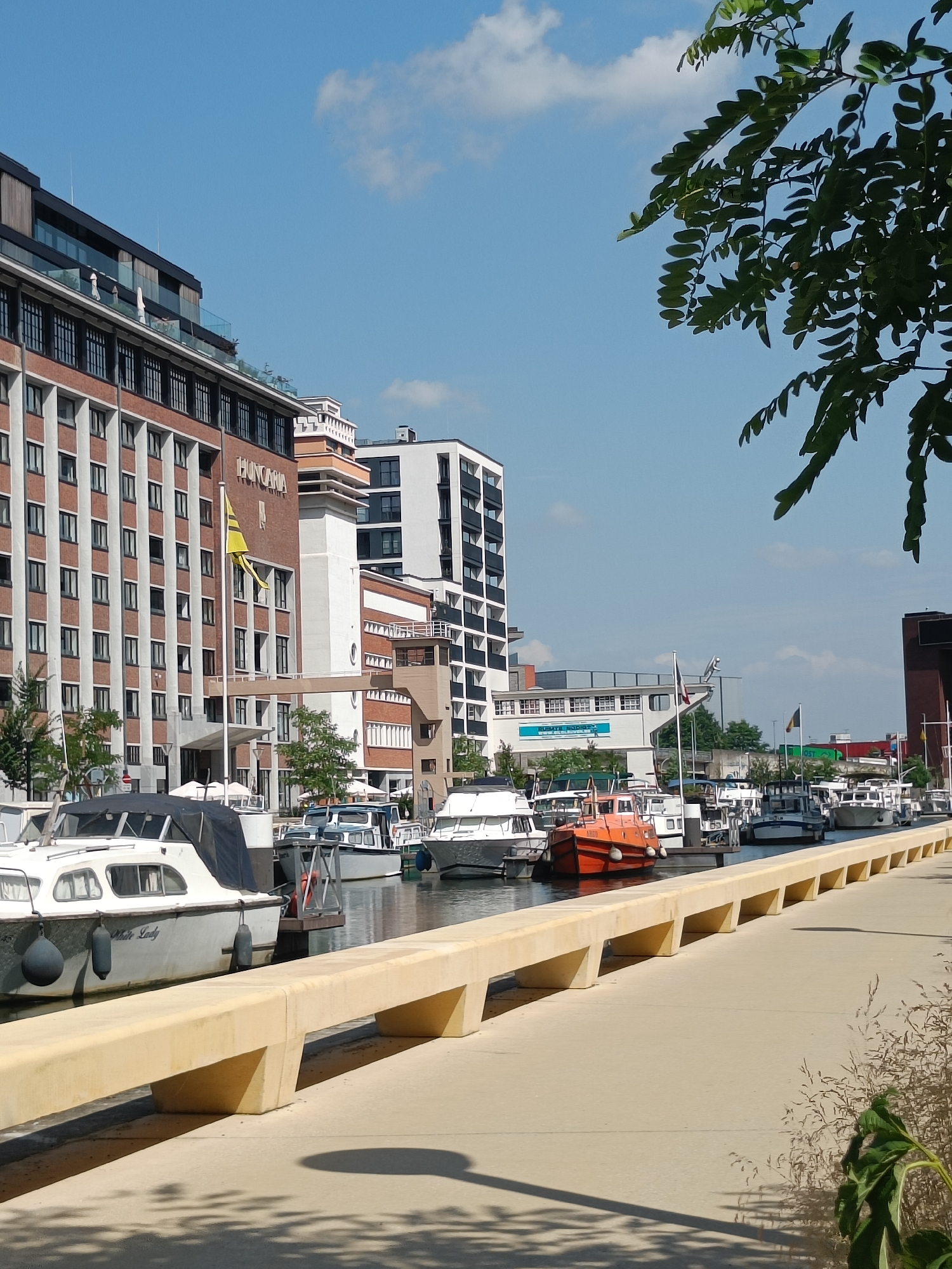 The image depicts a sunny day in a city canal, likely in Europe. The scene is dominated by a modern waterfront with several buildings and a row of docked boats. In the foreground, a light-colored concrete promenade runs along the canal, bordered by a yellow concrete bench. To the left, several boats are moored, including a white boat named "White Lady" with a dark canopy. Beyond the boats, a large brick building with numerous windows is visible, bearing the name "HUNGARIA" and a yellow flag. Further down the canal, more boats and modern buildings with glass facades can be seen. The sky is blue with scattered white clouds, and the branches of a tree with green leaves hang into the frame from the upper right corner, casting shadows on the promenade.