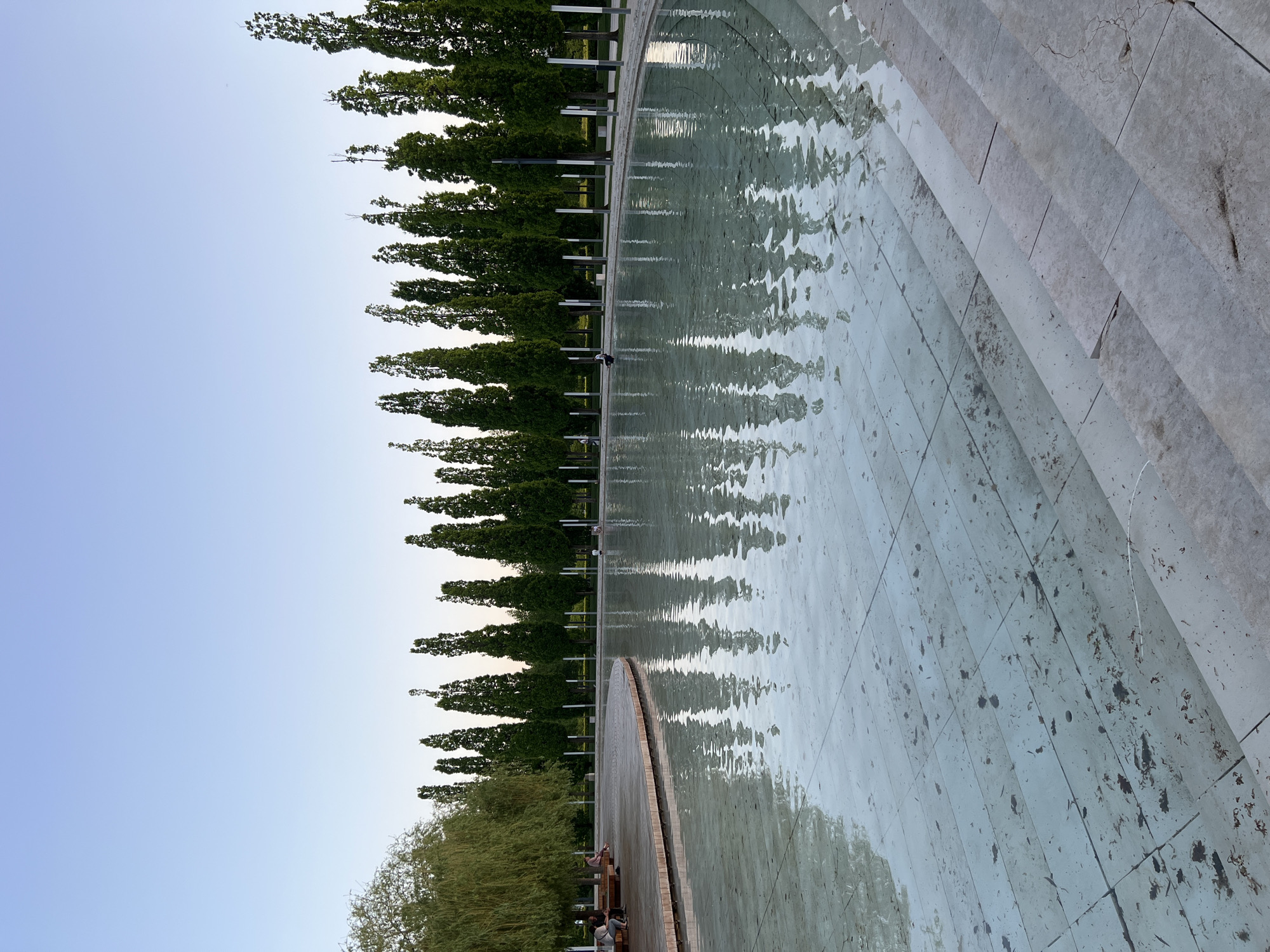 The image depicts a scene at the Galitsky Park in Krasnodar, Russia. A large, shallow, tiled pool with ripples covers much of the foreground, with steps on the right side leading into the water. Along the left side of the pool, a row of tall, thin trees line the edge. Their reflections distort on the water's surface. Further in the lower left corner are some other trees. Along the edge of the pool, on the left, there are some benches. Some people are sitting on these benches. The sky is a light blue, almost white, visible above the trees. Metal light poles are installed intermittently between the trees. The overall lighting suggests it is either early morning or late evening.