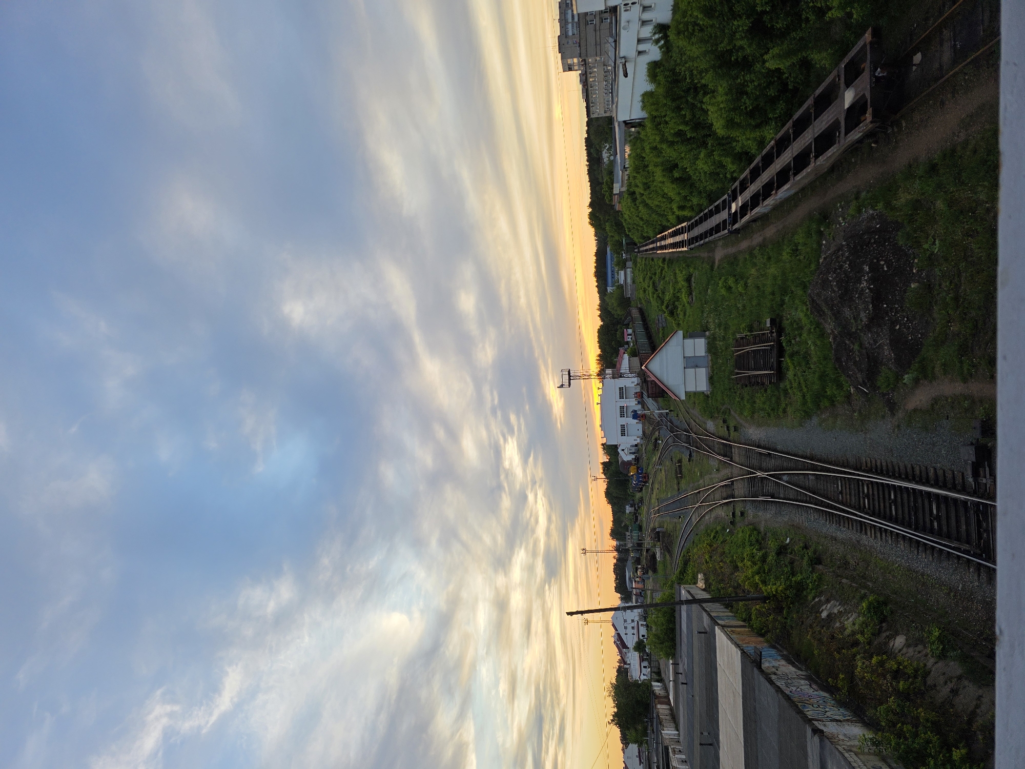 The image depicts a view from an elevated position, possibly a bridge or high building, overlooking a railway yard in what appears to be the city of Chaikovsky, Russia. The sky is filled with streaks of clouds illuminated by the setting sun, transitioning from blue to orange.
Below, a complex network of railroad tracks crisscrosses through the yard. Several buildings, including apartment blocks and industrial structures, are visible along the track's edge. Lush greenery punctuates the scene, with trees and grass growing beside the tracks and around the buildings.
In the background, a long row of railroad cars is positioned along the right side of the image. The buildings are predominantly white and grey, and the surrounding landscape features a mix of paved areas, vegetation, and gravel. A concrete structure is in the bottom right corner, presumably the base for the location of the photo.