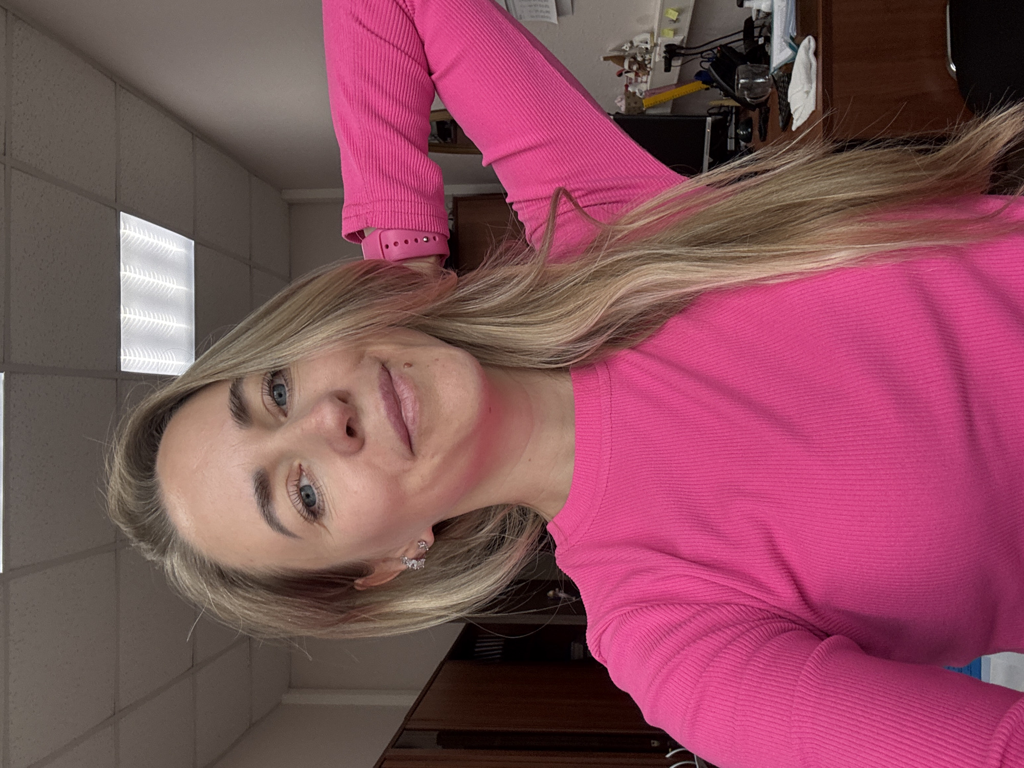 A woman with long, straight, blonde hair and light blue eyes is featured in a selfie, rotated 90 degrees counter-clockwise. She has light skin and a gentle, closed-mouth smile, looking directly at the camera. Her blonde hair is parted on the side and falls over her shoulders and chest. She is wearing a bright, hot pink ribbed long-sleeved top and a matching pink smartwatch on her left wrist. Small, sparkling earrings are visible on her ears, with a stud in her left earlobe and a small hoop in her right.
The background indicates an indoor setting, likely an office or institutional building in Chaikovsky, Russia. A drop ceiling with square tiles and a brightly lit fluorescent panel are visible above and behind her. Dark wooden furniture or cabinetry can be seen in the background, along with some indistinct items and clutter on a shelf or desk. The lighting is bright and even, characteristic of indoor fluorescent illumination. No discernible text is visible in the image.