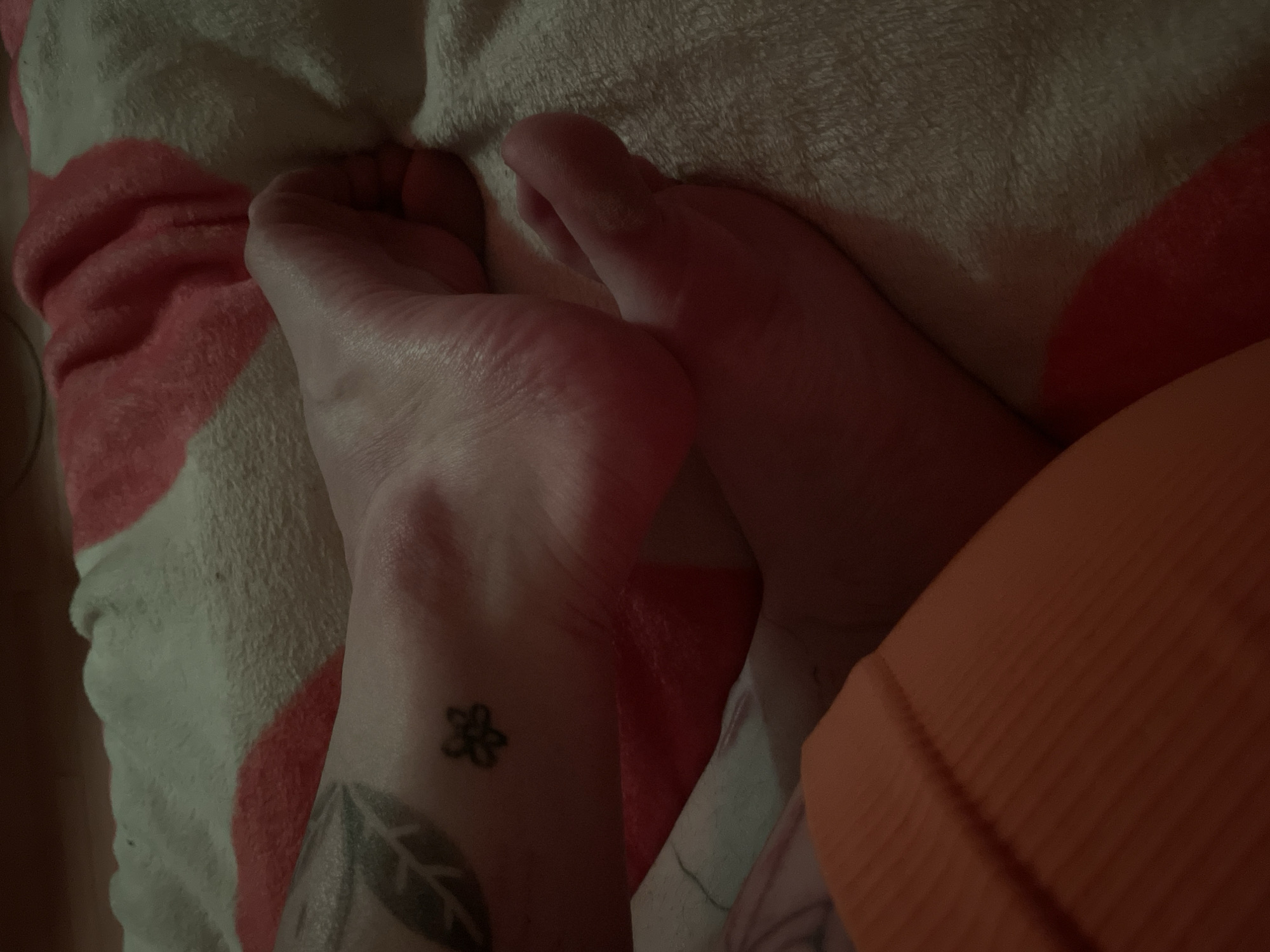 The image depicts two feet resting on a blanket with a red and white pattern. A small flower tattoo is visible on the ankle of the left foot. A larger leaf-shaped tattoo is partially visible below the flower tattoo. An orange fabric object is present on the right side of the image. There is no evidence to place this scene in Belgrade, Serbia.