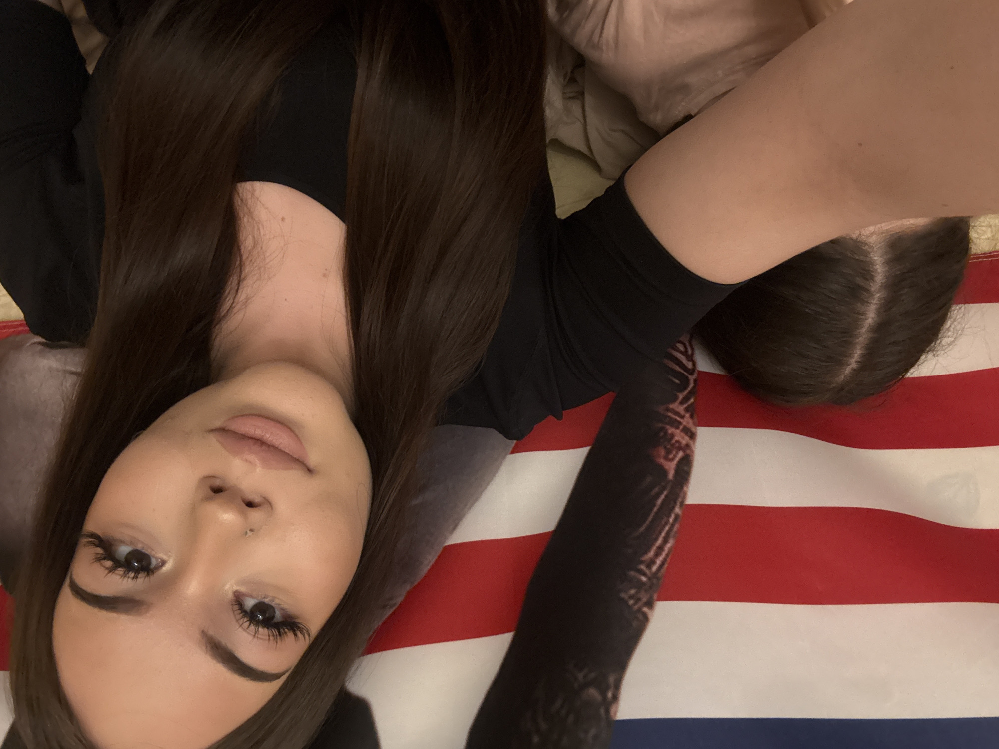 An indoor, eye-level portrait shows a young woman with long, dark brown hair lying down, looking directly at the camera. Her hair, parted on the side, frames her face and flows around her shoulders. She has fair skin, full lips, defined eyebrows, and extended eyelashes, along with a small piercing visible on her nose. She is wearing a black long-sleeved top; a dark, intricate pattern or possibly a tattoo is visible on her right sleeve. The woman is positioned on an American flag, which covers the surface beneath her, prominently displaying its red and white stripes and a small section of the blue field. Portions of a light-colored blanket or cushion are visible near her head and upper body. The lighting is warm and artificial, suggesting an indoor setting, likely in Naberezhnye Chelny, Russia. Her expression is neutral and calm. No text is visible in the image.