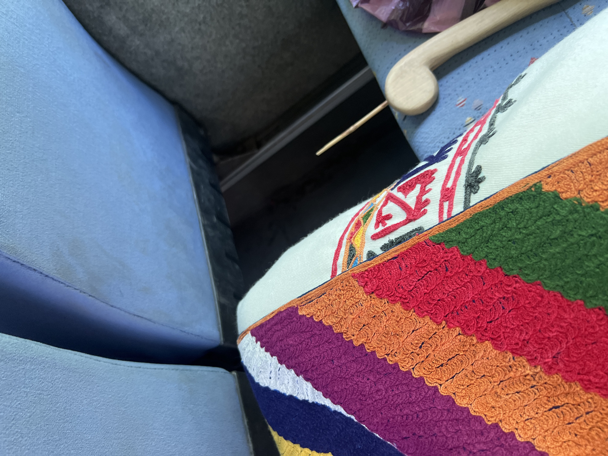 An interior scene captured in the city of Slivovo, Bulgaria, focuses on a person's lap. A white garment covers the lap, upon which a vibrant, multi-colored textile is resting. The textile features distinct diagonal stripes in dark blue, white, purple, orange, red, and green. The purple, orange, and red stripes are characterized by a pronounced textured, scalloped edge. The white section of the textile, towards the right, displays embroidered geometric patterns and stylized motifs, predominantly in red, orange, and dark grey. In the background, two light blue, textured vehicle seats with visible stitching are partially visible, separated by a dark, recessed area. To the immediate right of the textile, a light-colored wooden object, resembling the top portion of a coat hanger, rests on a blue surface that exhibits faint patterned details. Behind the wooden object, a crumpled, reddish-purple piece of packaging is visible. No individuals are fully depicted; only the implied presence of the person whose lap is shown.