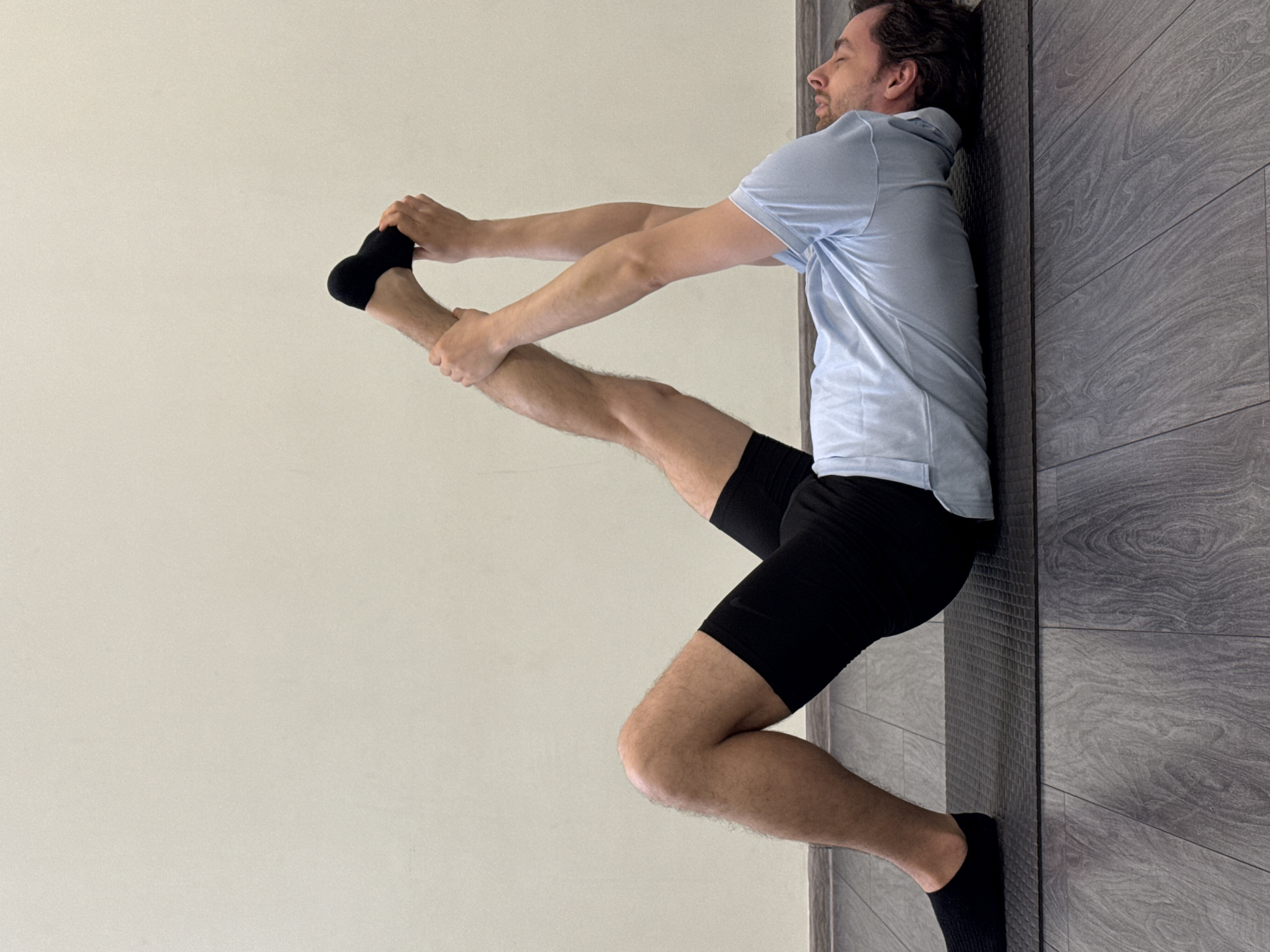 A man in black shorts and a light blue shirt is stretching against a wall. He is standing on his left leg, which is bent at the knee, and his left foot is on the floor. His right leg is extended in front of him, and he is holding his right foot with both hands. The man is facing to the left. The wall behind him is a dark gray, and the wall in front of him is a light beige. The location is Unknown, Unknown.