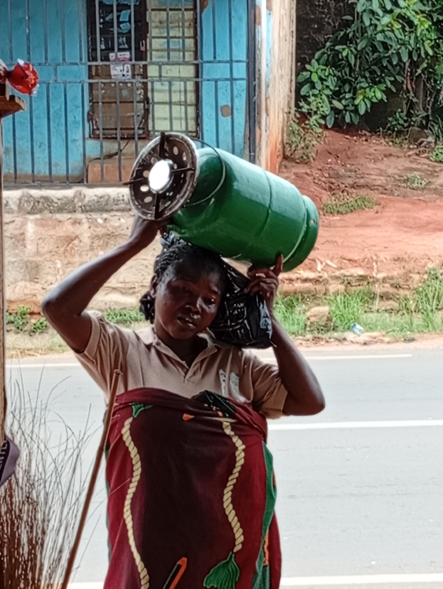 A woman carries a green gas cylinder on her head in an outdoor setting in an unknown city. The cylinder has a black circular component attached. The woman is wearing a tan collared shirt and a maroon and green patterned cloth. She also carries a black plastic bag on her shoulder. In the background, there is a building with a blue-painted barred window. To the right, there is green vegetation. A grey road is visible in the foreground.