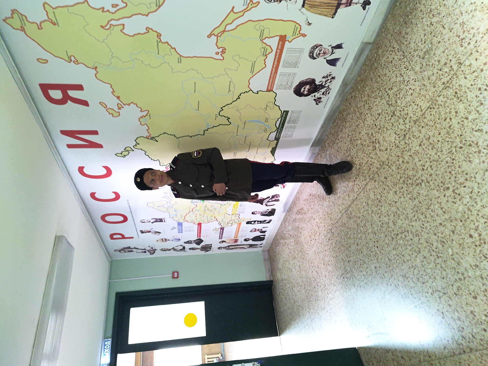A man in a dark green military-style uniform and black boots stands at attention in what appears to be a hallway. He is wearing a dark cap. Behind him is a large wall map labeled "Россия" (Russia) in large red letters, with various images of people associated with the regions within Russia. The hallway has a tiled floor and an exit door with a window. The location is likely Markovskii, Russia, considering the text and map in the background.