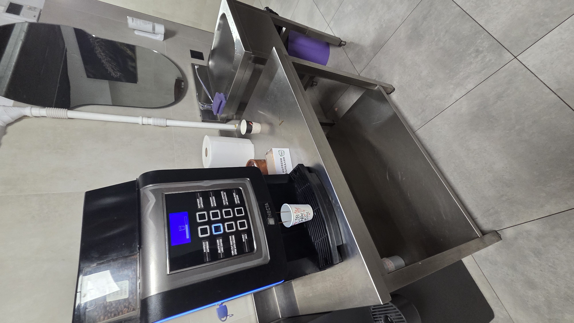 A modern automatic coffee machine is positioned on a stainless steel counter in a clean, commercial-style kitchen or breakroom. The machine features a digital display and multiple buttons, with a cup actively being filled with coffee. Next to the coffee machine is a roll of paper towels, a small sink with a faucet, and a stainless steel sink basin. Above the sink, a large, oval mirror reflects the room, showing a framed picture and a white cleaning tool. The floor is tiled in a neutral gray color. There are no people visible in the image. The scene appears to be an operational coffee station.
