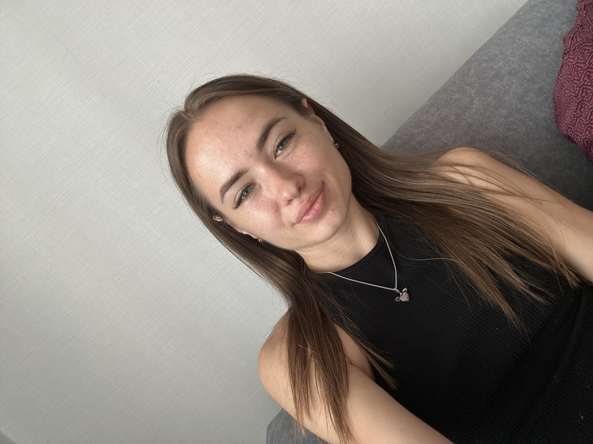 In the center of the frame, a young woman with long brown hair is captured in a selfie, her head tilted slightly. She wears a black sleeveless top and a silver necklace with a heart-shaped pendant. Her gaze is directed towards the camera, and her lips are curved in a soft smile. The background consists of a textured grey wall to the left, a grey sofa in the center, and a hint of a patterned purple cushion on the right. The lighting appears to be natural, casting a soft glow on her face.