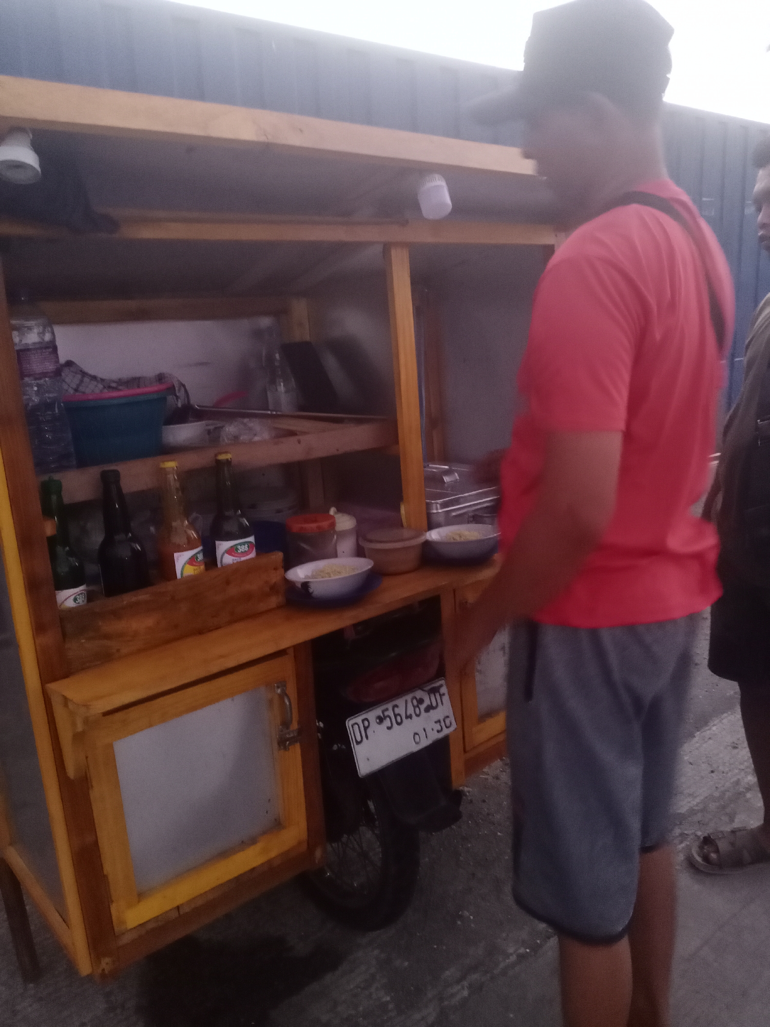 The image depicts a food cart in Siddo, Indonesia. A man wearing a red t-shirt and gray shorts stands by the cart, which is made of wood and contains various items for sale. These include bottles of condiments, bowls of food, and a lightbulb attached to the top shelf. A motorcycle license plate is visible on the side of the cart. Another person is partially visible on the right side of the image. The setting appears to be an outdoor location, possibly a street or market.