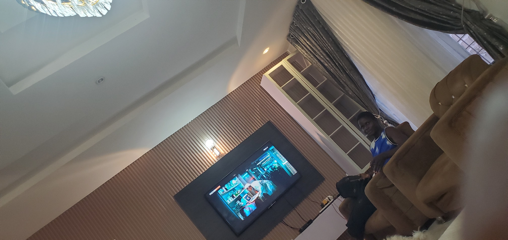 The image depicts an indoor scene with a modern living room setup. A large television screen mounted on a textured brown wall displays a scene from what appears to be a science-fiction or medical drama, featuring people in lab coats. The television is positioned above a dark entertainment unit.
In the foreground, two individuals are seated on plush, tufted brown sofas. One person, partially obscured, is lying down on the sofa. The other person, a young man wearing a blue and white patterned tank top, is sitting upright, looking towards the camera with a slight smile.
Behind the sofas, a tall, white shelving unit with multiple compartments is visible. Dark grey curtains are drawn across a window or doorway on the right side of the frame, behind the seating area. The ceiling is white and features recessed lighting and a decorative chandelier on the upper right side of the image. The overall impression is that of a comfortable and contemporary living space.
