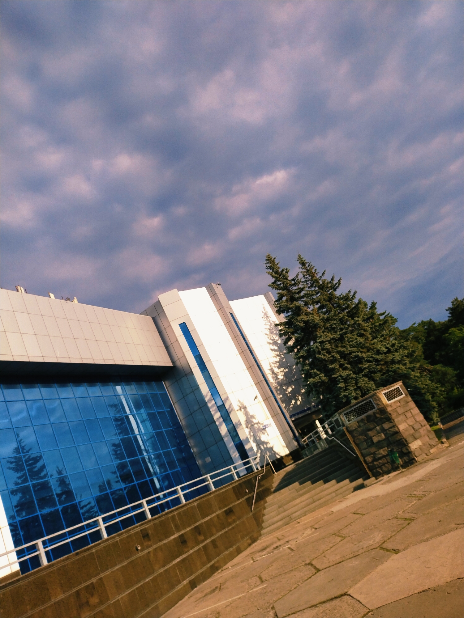The image depicts a modern building with large blue glass windows, a white facade, and metallic accents. The building is situated in Horishni Plavni, Ukraine. In front of the building, there are stone stairs leading up to an entrance. To the right of the stairs is a stone structure with a grated opening. Evergreen trees are visible behind the building, and a cloudy sky fills the background. The foreground consists of a paved walkway with large stone slabs. The scene appears to be outdoors during the day, with the sun casting shadows on the building and walkway. There are no people visible in the image. The overall impression is one of an urban landscape with architectural and natural elements.