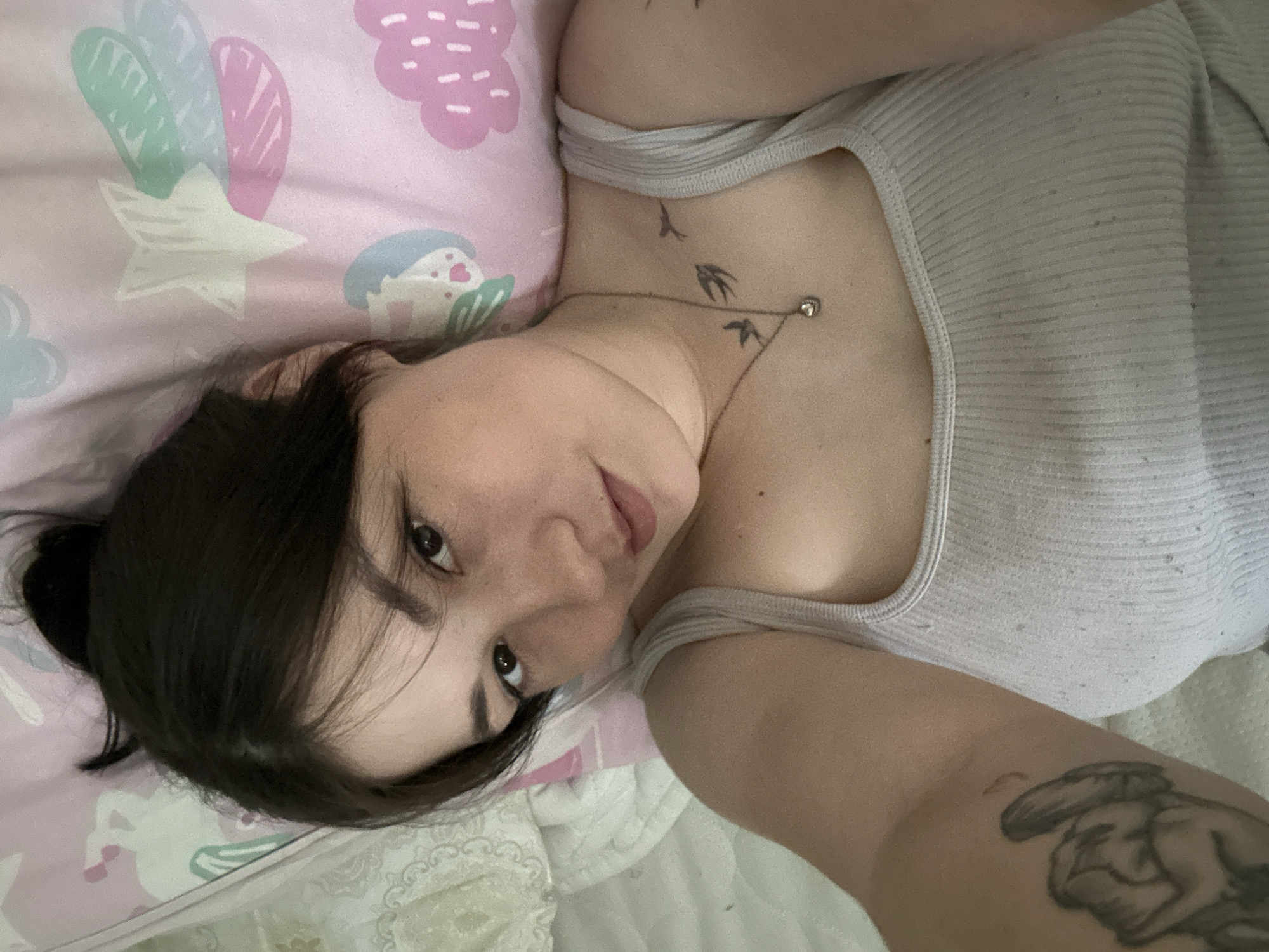 A young woman with dark hair pulled into a bun is lying on her side, looking at the camera. She is wearing a light gray tank top and a silver necklace with a small circular pendant. There are tattoos on her arm and collarbone. The background is a pink pillow with a pattern of stars and clouds. The scene appears to be in a bedroom. There is no indication of the location within the city of Chaikovsky, Russia.