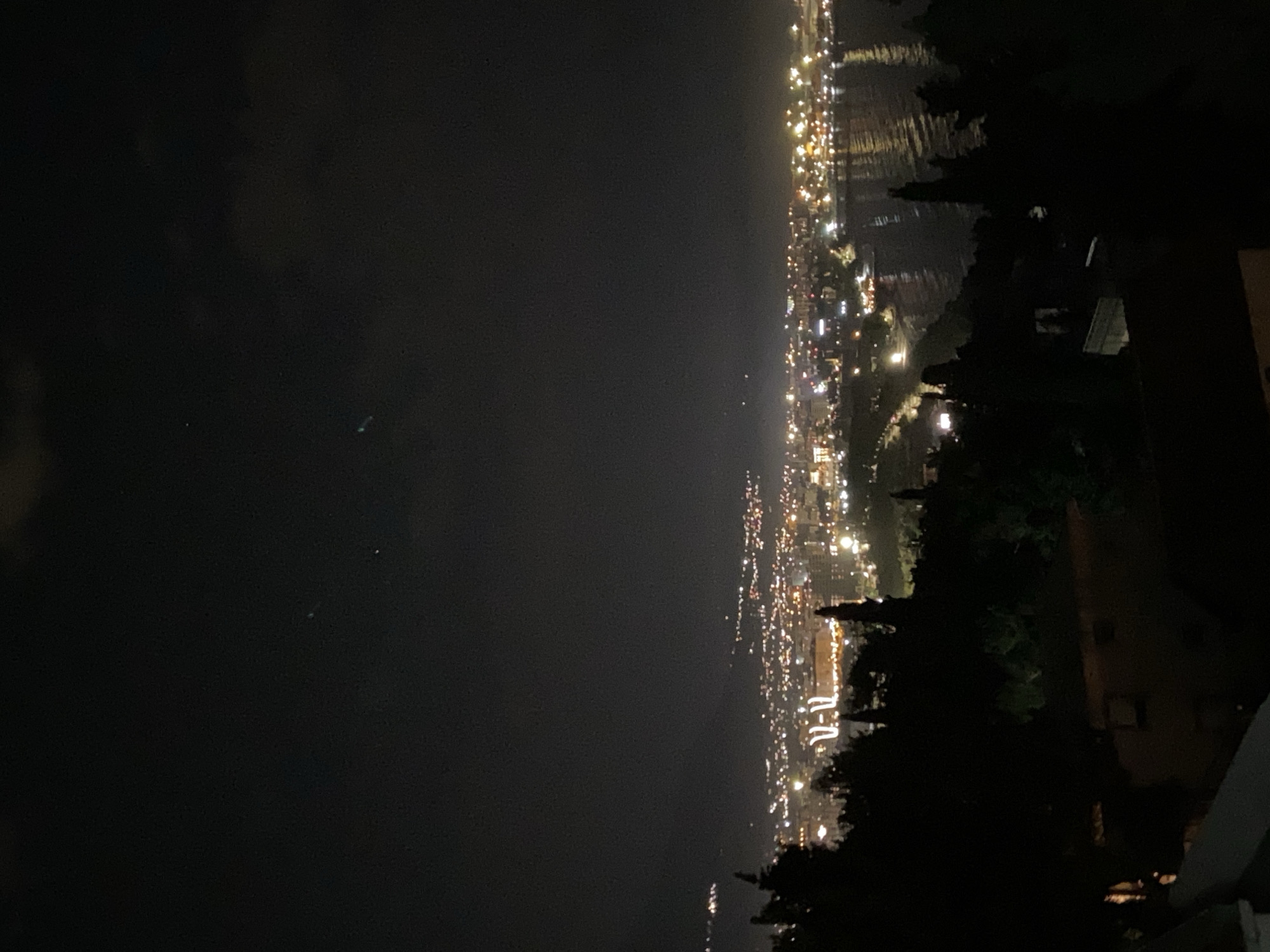 The image depicts a nighttime view of the city of Šušanj, Montenegro. The city lights are visible along the coastline and extend inland, reflecting on the water. In the foreground, dark shapes of trees and a building obscure part of the view, indicating the vantage point is elevated, possibly a hillside. The sky is dark with some cloud cover.