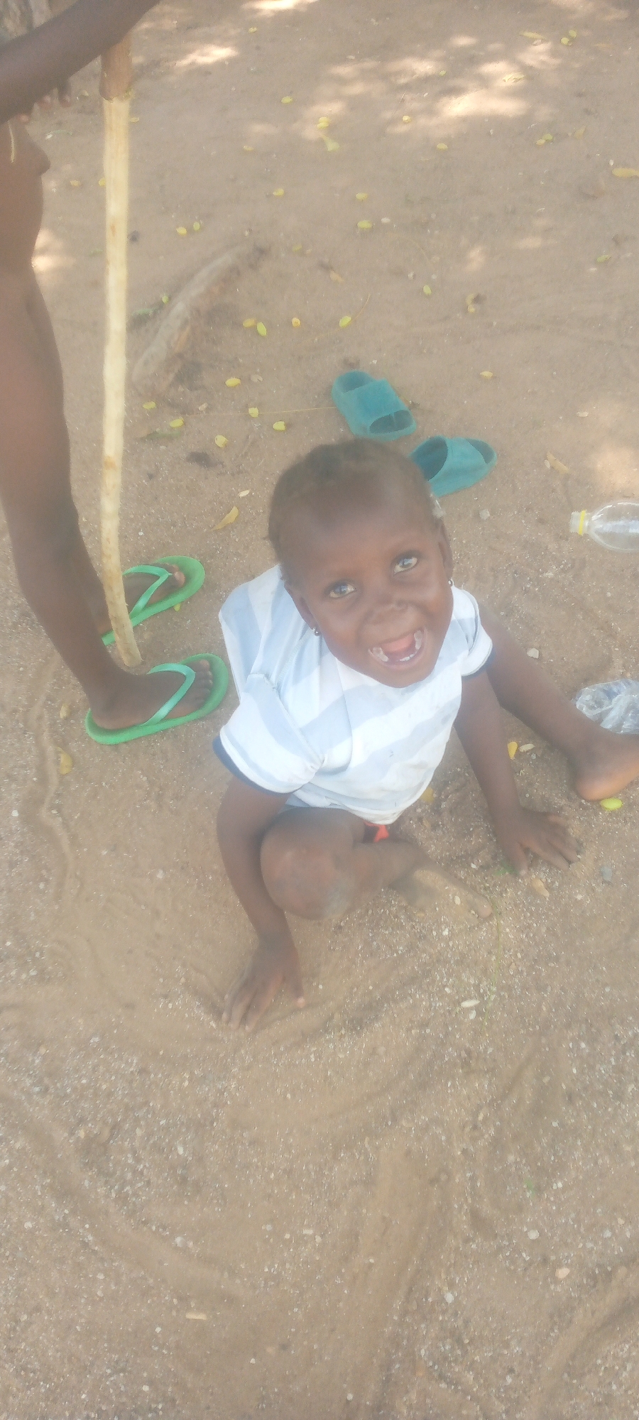 A young child, possibly a toddler, is sitting on sandy ground, looking up at the camera with an open mouth and wide eyes. The child is wearing a white t-shirt with thin blue stripes and an orange garment underneath. To the left of the child, a person's leg and foot are visible, wearing bright green flip-flops. This person is holding a light-colored wooden stick. In the background, two teal-colored flip-flops are visible on the sand. The ground is covered in sand and small pebbles, with scattered yellow seed pods or leaves. Sunlight casts dappled shadows from overhead foliage onto the scene. A partially visible plastic water bottle is also present on the right side of the frame.