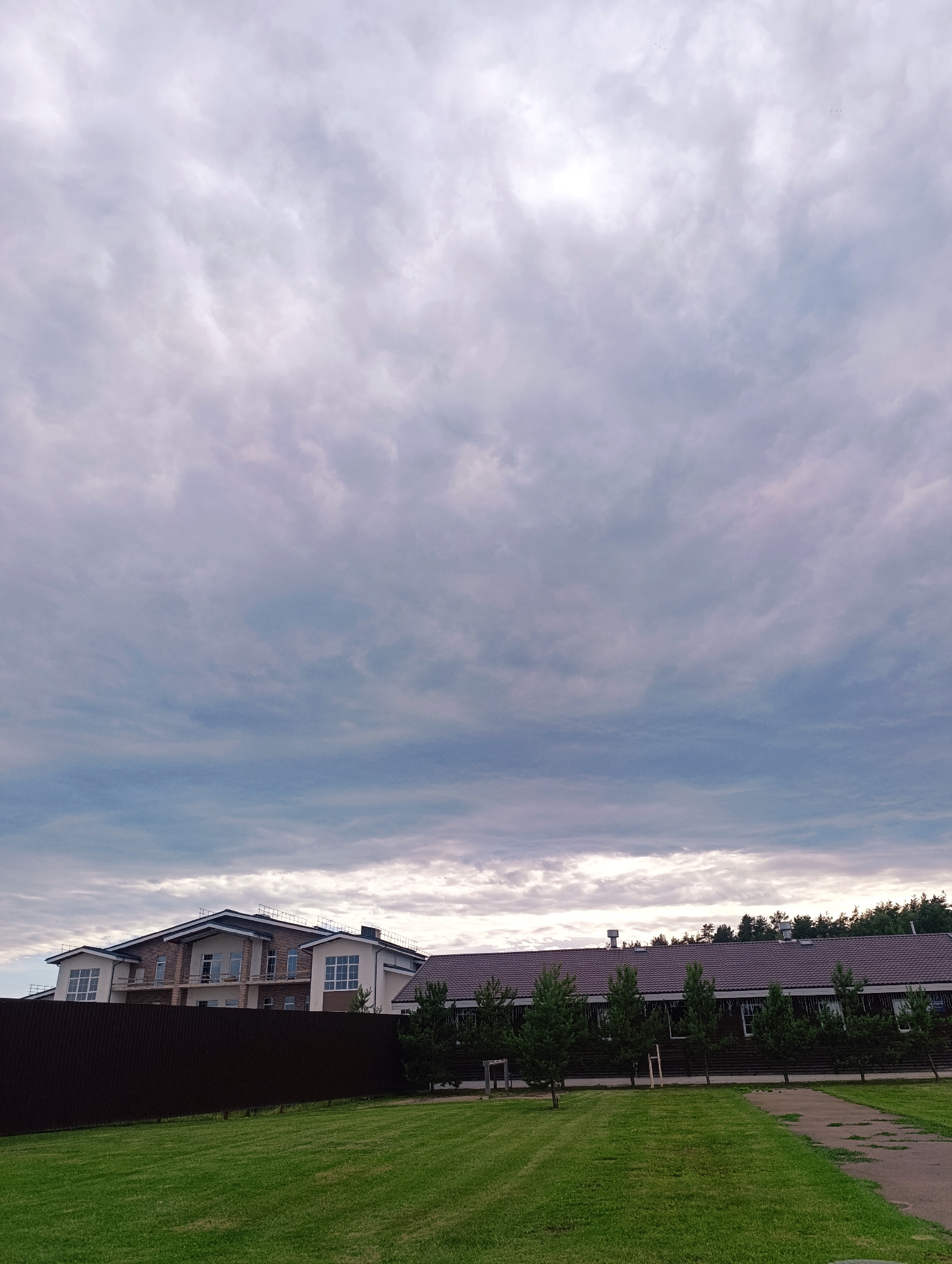 This media file depicts an outdoor scene under a cloudy sky. The foreground features a well-maintained green lawn with a paved pathway on the right. In the mid-ground, a dark brown, corrugated metal fence runs horizontally across the image, separating the lawn from the properties behind it.
Beyond the fence, two buildings are visible. The building on the left is a large, multi-story house under construction, made of brick and stucco, with visible windows and balconies. To the right of this house is a long, low-rise building with a dark brown tiled roof and numerous windows. A line of small evergreen trees stands in front of this building, and some lights, possibly for decoration, are strung along the eaves. A dense line of trees forms the background, suggesting a wooded area.
The sky is filled with dramatic, heavy clouds in shades of gray and white, indicating overcast or impending rain weather. The lighting is diffused, suggesting it is either early morning or late afternoon, but the overall atmosphere is somewhat subdued due to the cloudy conditions. There are no people visible in the image, and no text is present. The scene suggests a residential or possibly a recreational area in a rural or suburban setting, with the construction on one house indicating ongoing development. Given the location context of Ivanovskoe, Russia, this could be a view of a private property or a community area within that region.
