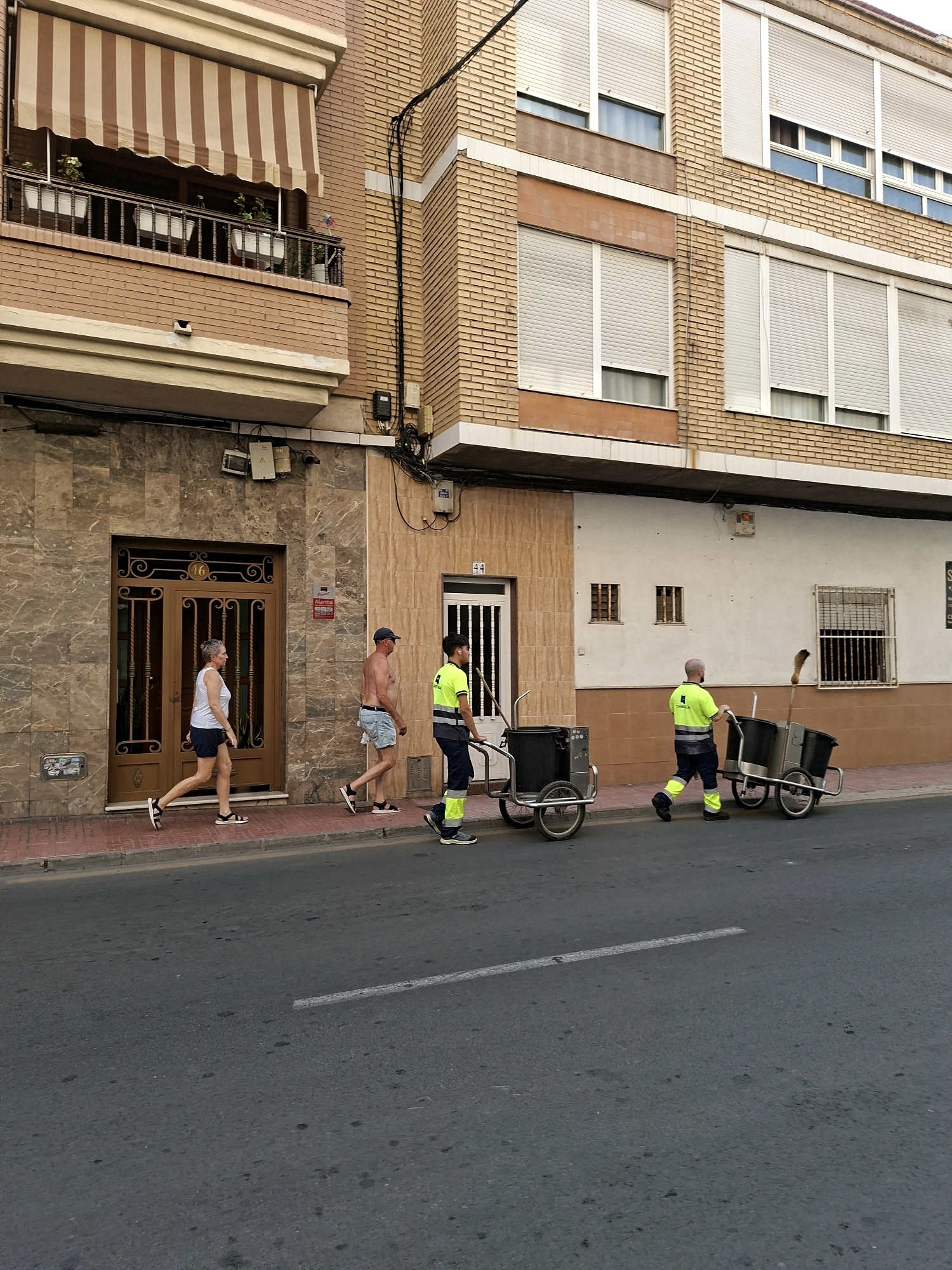 The image depicts a street scene in Torrevieja, Spain, likely in the daytime, with a focus on the sidewalk and the adjacent building. In the foreground, a paved street with a white line marking is visible. On the sidewalk, three individuals are walking. The first person from the left is a woman with short gray hair, wearing a white tank top and dark shorts, and sandals. She is walking towards the right. Next to her, a shirtless man with a blue baseball cap and denim shorts is also walking to the right. Following them is a man wearing a high-visibility yellow jacket with reflective stripes and dark pants, pushing a cart with a broom. Another person in a similar high-visibility jacket and dark pants is partially visible on the far right, also pushing a cart. The carts appear to be waste collection bins on wheels. The building behind them is a multi-story brick structure with several windows covered by roller shutters. The ground floor features a large decorative doorway and a smaller entrance with the number "44" above it. The overall scene suggests a routine day with city workers performing their duties.
