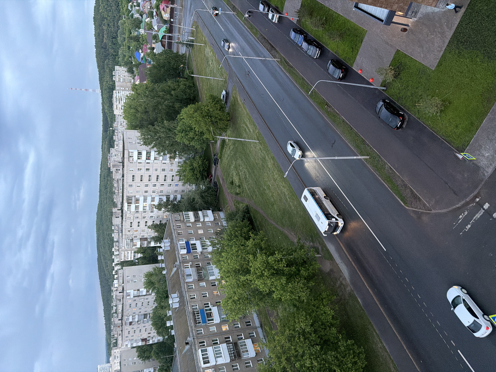 An aerial view captures a cityscape under an overcast sky in Unknown, Unknown. In the distance, a red and white tower rises above a line of trees. Several apartment buildings with rows of windows extend into the middle ground, bordered by mature trees. A road cuts through the scene, featuring multiple vehicles including a white bus and several cars. Streetlights are spaced along the road, illuminating the pathway. Patches of green grass line the road. A crosswalk symbol is visible on the roadside.