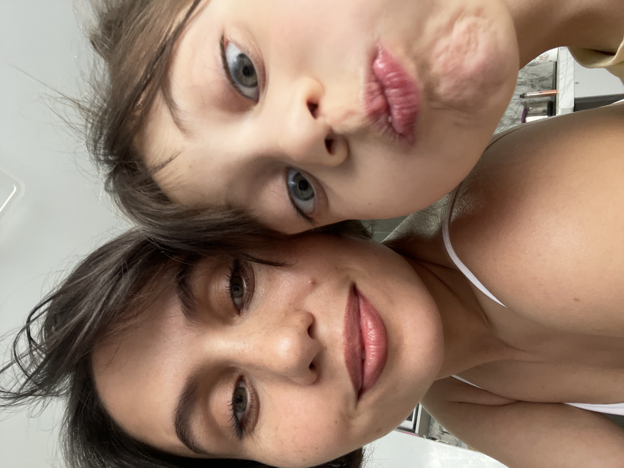 A close-up selfie shows a woman and a child making faces at the camera. The child is on top, puckering their lips and looking up. The woman is below, smiling slightly with a light lipstick. Both have dark hair. The background is out of focus and nondescript. The location is not identifiable as Izhevsk, Russia from this image.
