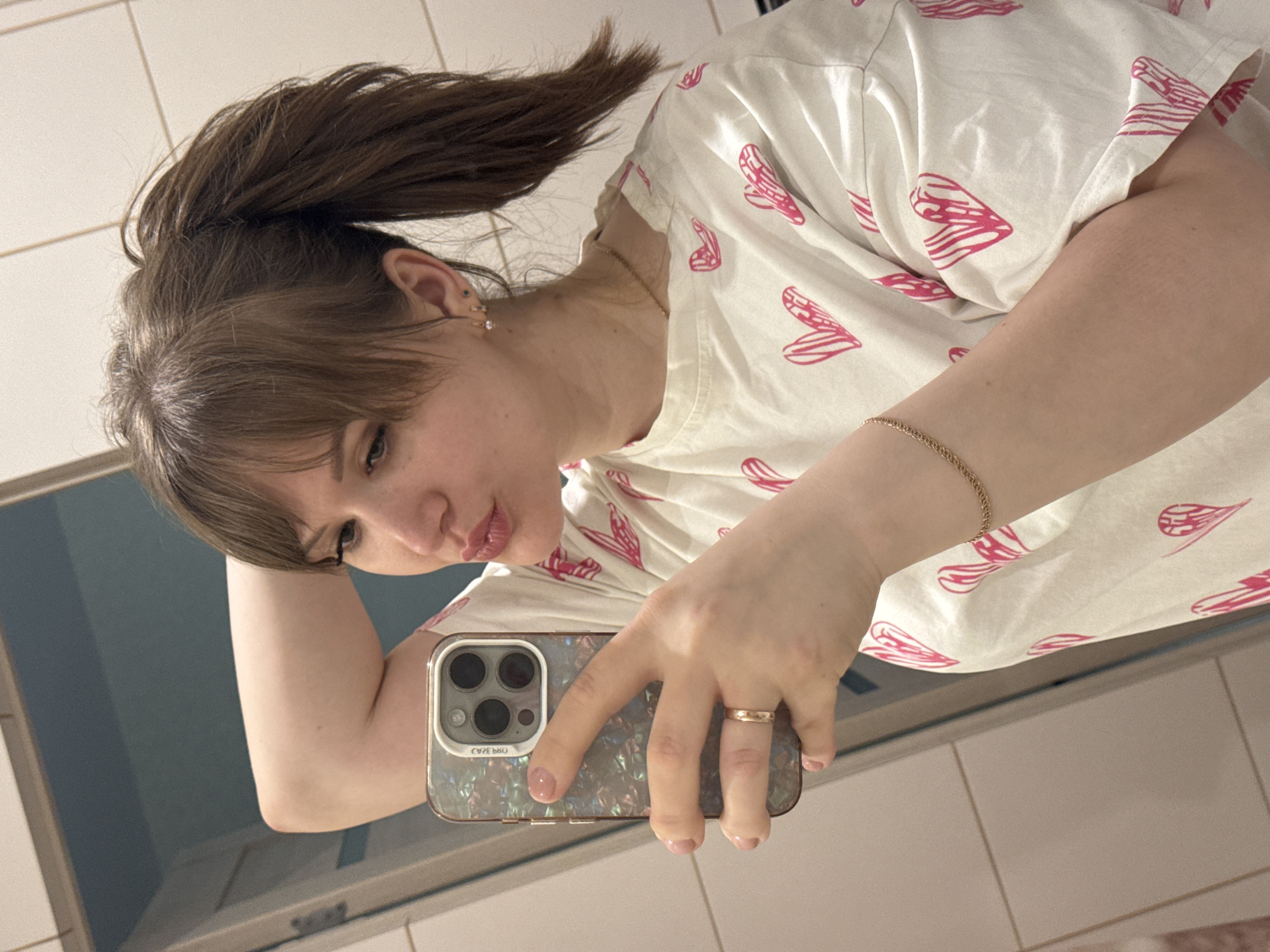 A woman with brown hair in a high ponytail is taking a selfie in a bathroom. She's wearing a white t-shirt with pink heart-shaped designs. Her left arm is bent, holding her phone, and she has a gold bracelet on her wrist. A gold ring is visible on her finger. The background shows a bathroom mirror and tiled walls. The location appears to be a residential bathroom. There is no specific information indicating that this image was taken in Saratov, Russia.