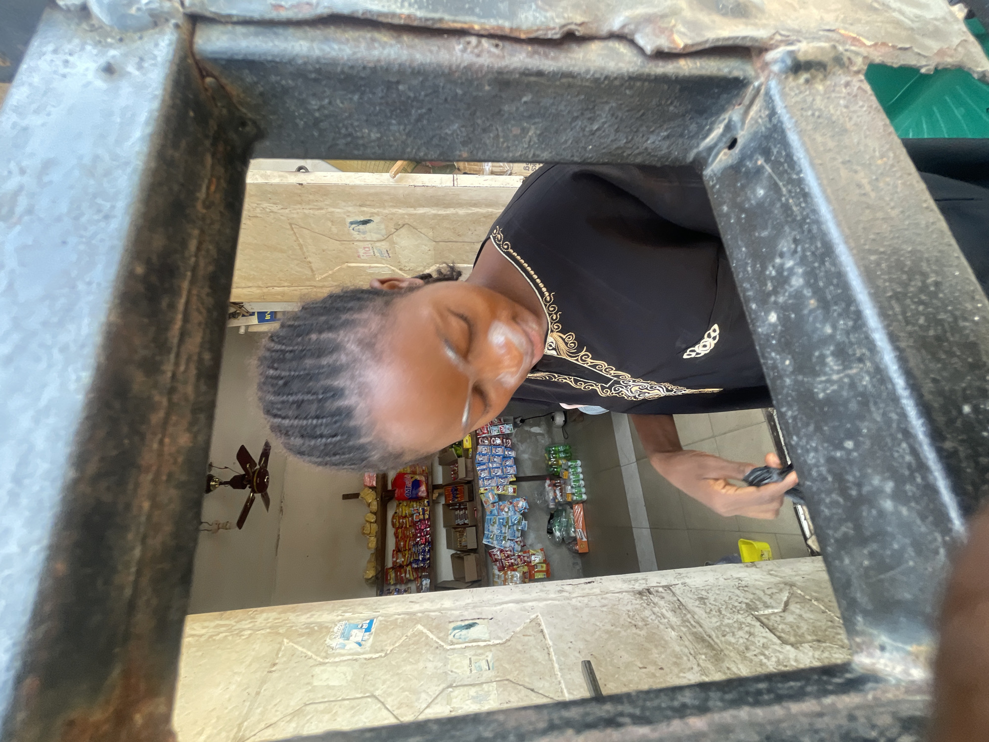 A woman with dark hair braided close to her head leans forward through a rectangular opening framed by a dark metal structure. She wears a black top with gold embroidery around the neck. Her eyes are closed. She holds a dark object in her hand. The opening looks into a small shop stocked with various goods on shelves. A ceiling fan is visible in the upper left. The scene is possibly in Lokoja, Nigeria.