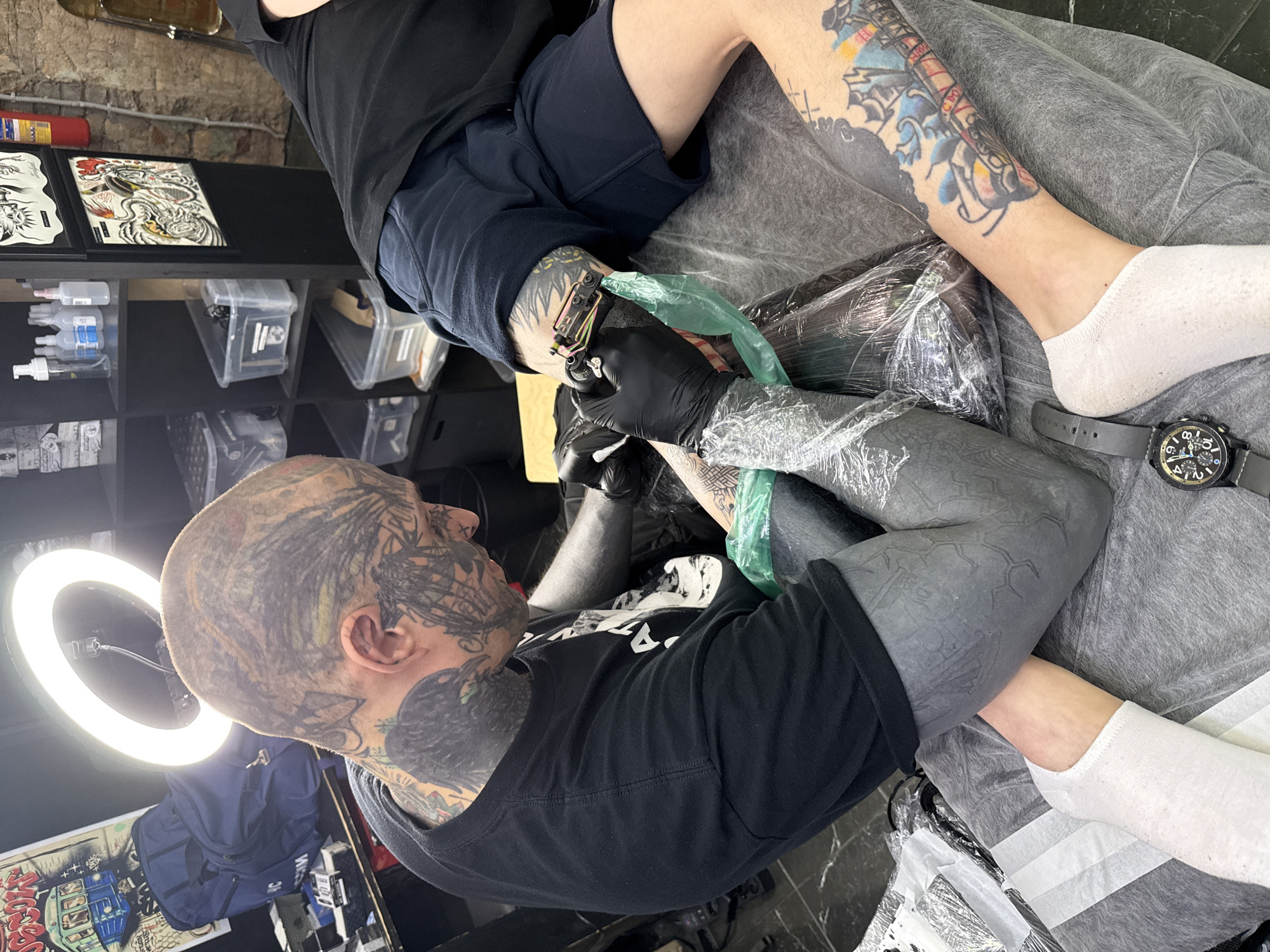 The photograph captures a tattoo artist working on a client's leg in what appears to be a tattoo studio. The artist, extensively tattooed on the head, neck, and arms, is wearing a black t-shirt and black gloves. They are focused on the client's leg, which is partially covered with plastic wrap, using a tattoo machine with a green covering. The client is wearing black shorts and white socks, with existing tattoos visible on the other leg. A watch is visible on the client’s wrist. In the background, shelves are stocked with tattoo-related supplies, and framed tattoo designs are displayed on a counter. A ring light is positioned to illuminate the workspace. The location is not identifiable as Moscow, Russia, based on the visual information.