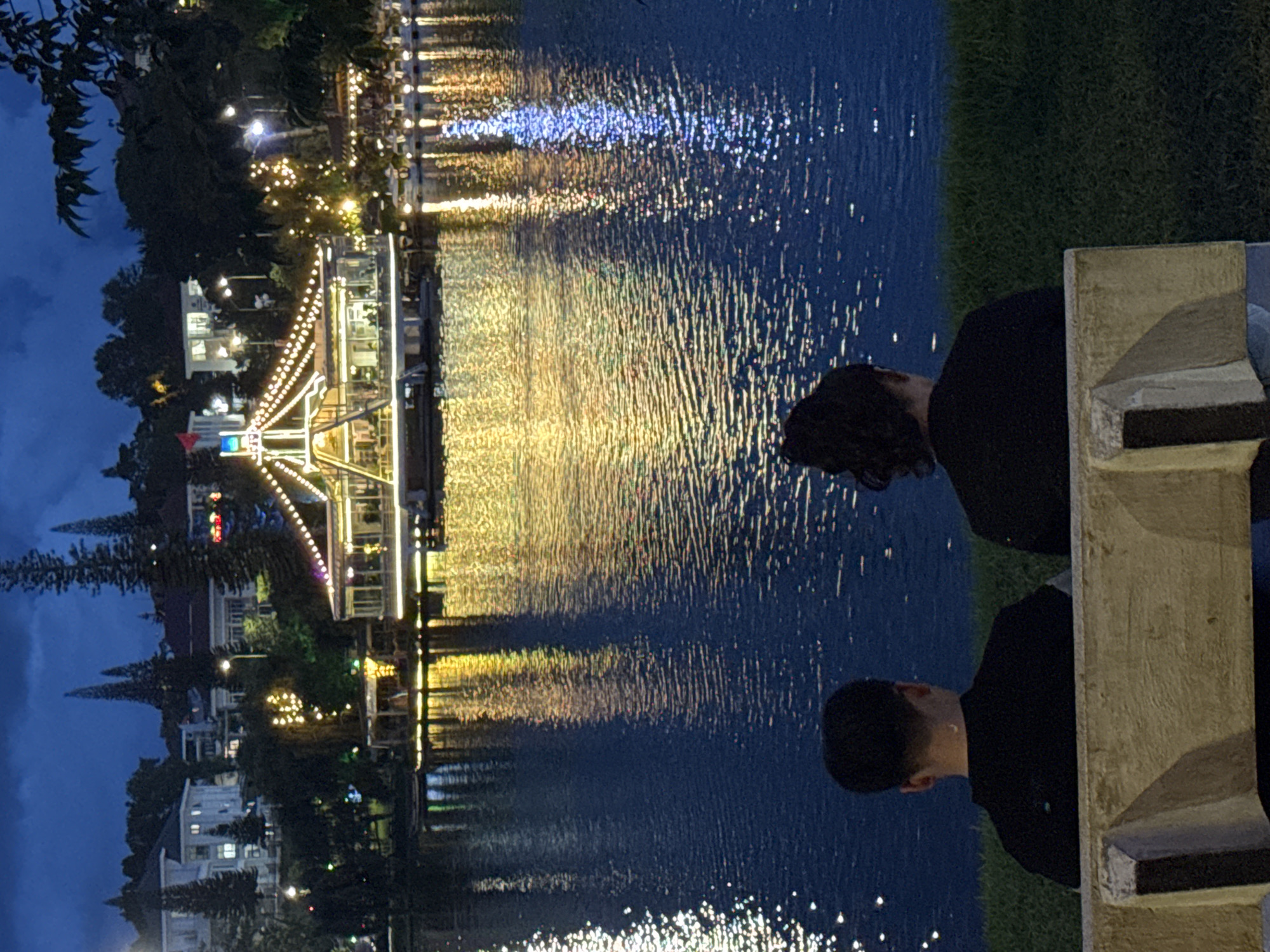 In Xuan Huong Lake, Da Lat, Vietnam, two individuals are seated on a concrete bench facing the water. The scene depicts a nighttime setting with the lake reflecting the illuminated buildings and decorative lights from a structure across the water. The structure, possibly a restaurant or entertainment venue, is adorned with strings of golden lights, and further lights are visible on the adjacent buildings and trees on the hillside. The water's surface is disturbed by ripples, causing the reflections to appear elongated and shimmering. The foreground includes a grassy area and the bench on which the two individuals are seated. Their backs are to the camera, so their features are not visible.