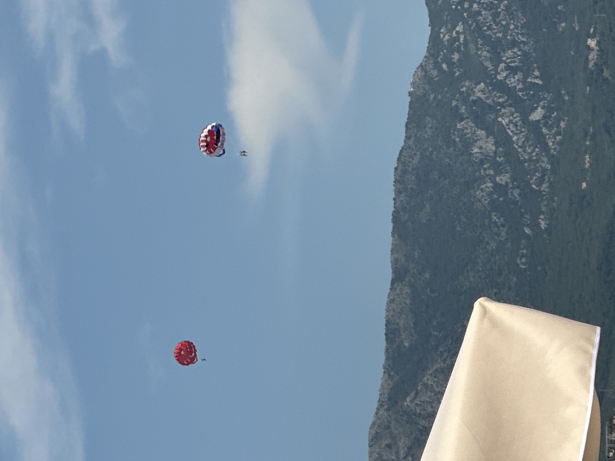 Two parasails are visible in the sky above a mountainous coastline in Budva, Montenegro. The parasail on the left is red and appears to be carrying one person. The parasail on the right is red, white, and blue and appears to be carrying two people. The coastline is covered in dark green vegetation and rocky outcrops. A large, cream-colored umbrella or canopy is in the foreground, partially obscuring the view of the coastline. The sky is blue with scattered white clouds.