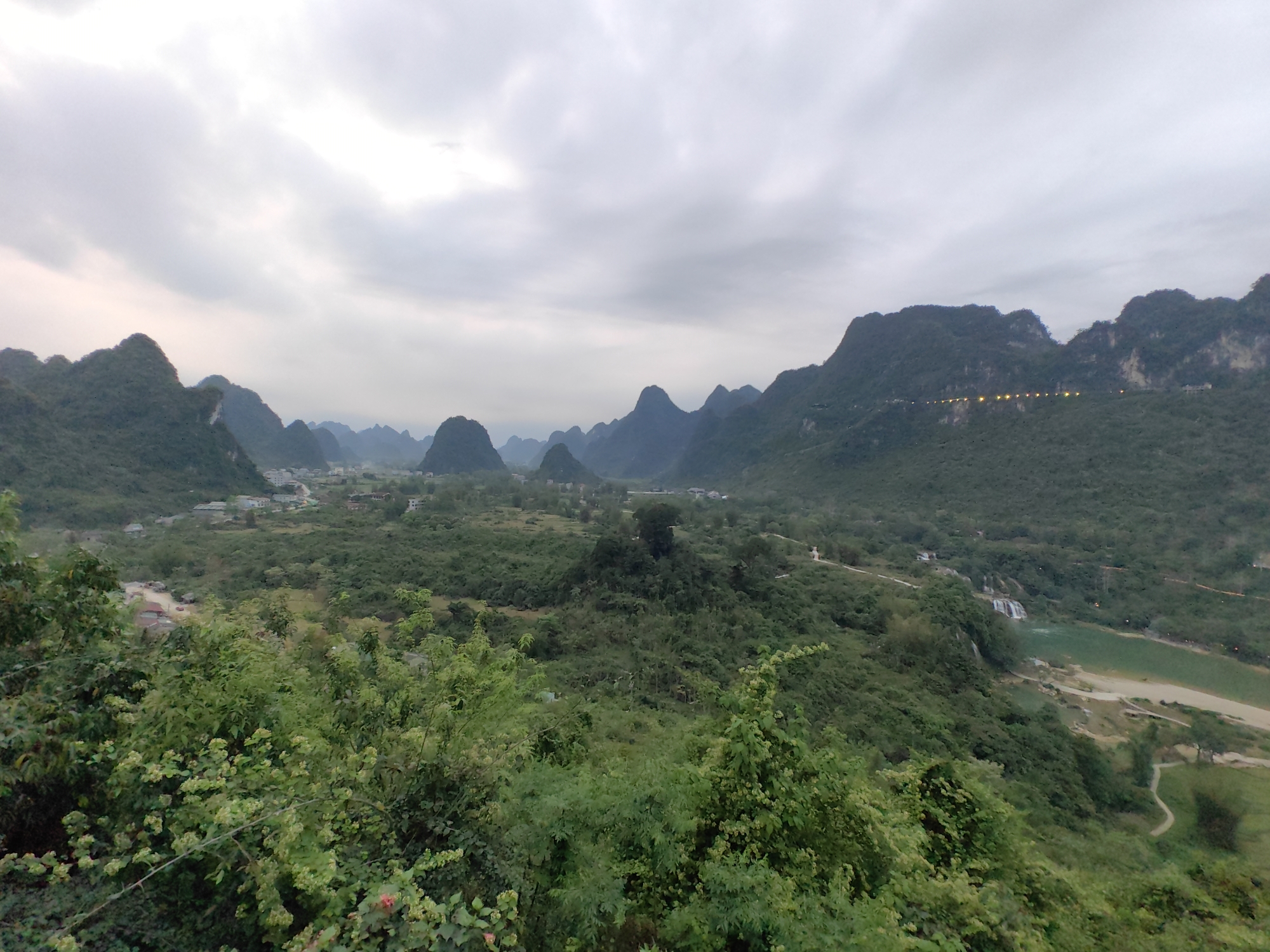 The image shows a panoramic view of the landscape in Xã Đàm Thủy, Vietnam. In the foreground, there is lush green vegetation. Beyond the vegetation is a valley with scattered buildings and structures, suggesting a small town. The valley is surrounded by prominent karst mountains with rounded peaks. On the right side, a road with lights is visible along the side of one of the mountains. A river or lake with a waterfall and a sandy shore can be seen towards the right of the image. The sky above is overcast with grey clouds, giving the scene a muted, somewhat hazy appearance.