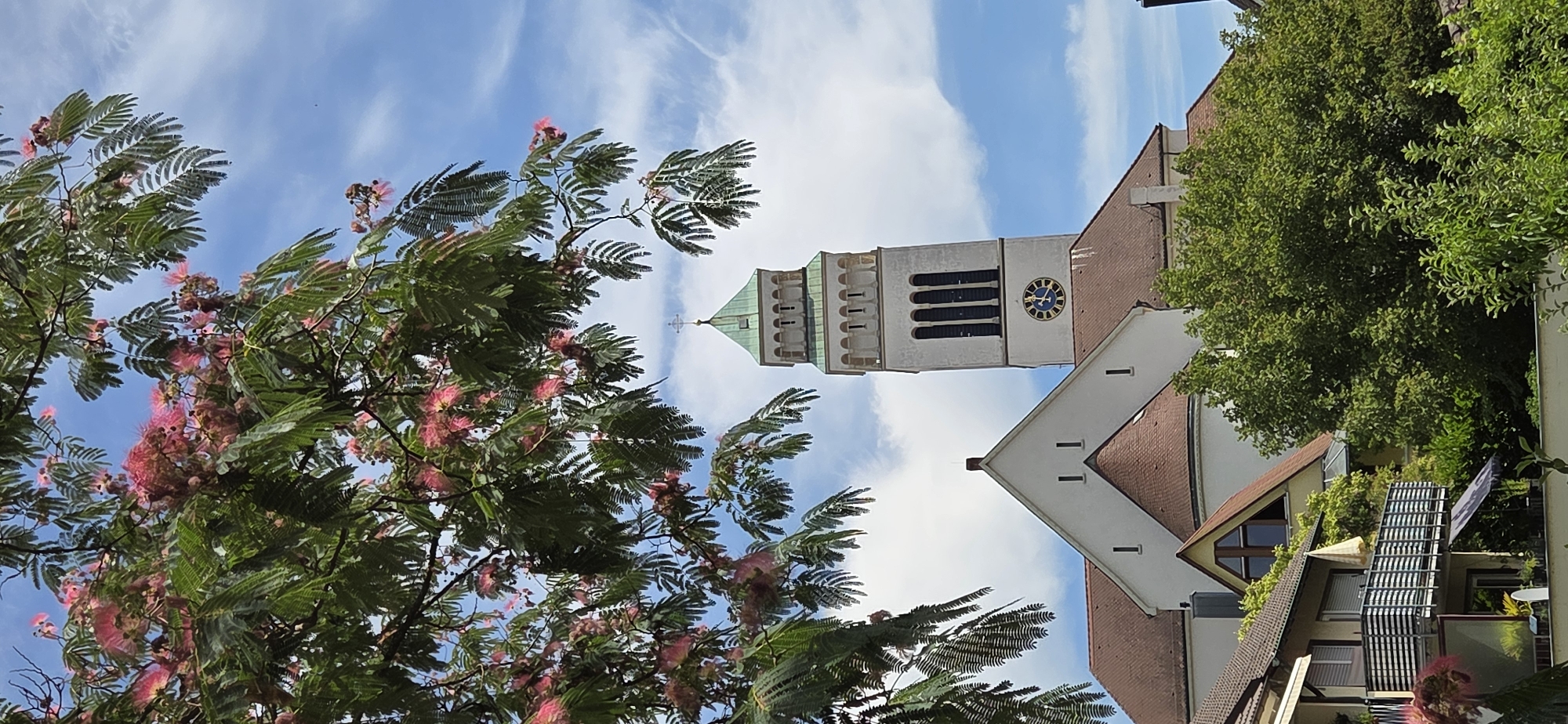 In Kehl, Germany, a tall white church tower with a green, pyramidal roof dominates the skyline. The tower features arched windows and a prominent clock face with blue numerals. Below the tower, the church has a white facade and a reddish-brown tiled roof, with a smaller conical roof section. In the foreground, lush green trees and branches laden with pink blossoms frame the scene. Residential buildings with balconies and windows are visible to the left and below the church, suggesting a mixed urban environment. The sky is blue with scattered white clouds.