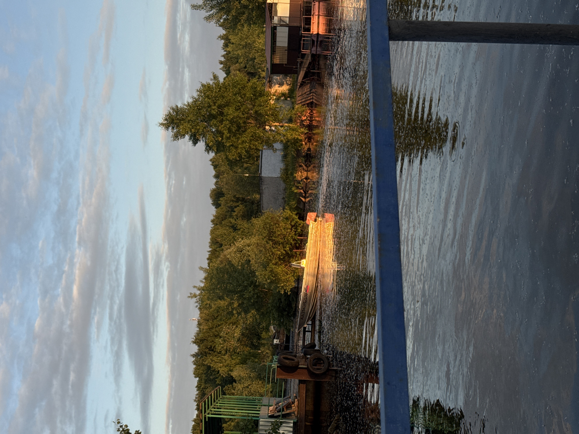 This image captures a tranquil riparian scene in Chaikovsky, Perm Krai, Russia, likely during either sunrise or sunset given the warm, low-angle sunlight. The perspective is from an elevated position, looking across a body of water, possibly a river or canal, towards the opposite bank.
The sky occupies the upper third of the frame, displaying a light blue hue with scattered, elongated grey and white stratocumulus clouds. The horizon is dominated by a dense line of various green deciduous trees, suggesting a forested area. A slender, white and red cellular communication tower rises above the tree line in the central distance.
The body of water in the middle ground and foreground is dark, reflecting the sky and the surrounding foliage, with subtle ripples on its surface. Patches of dark organic debris, possibly leaves or pollen, are visible floating on the water, particularly near the banks and in the foreground.
On the left bank, a green-framed structure with a metal roof, possibly a boathouse or covered dock, stands adjacent to a wooden pier or platform. Several black rubber tires are affixed to the side of this structure, serving as fenders. Moored to the pier is a light-colored aluminum boat, strongly illuminated by the golden sunlight, which reflects brightly off its hull. The boat displays the registration number "AA 2543 RUS 59" on its side, confirming its Russian registration from the Perm Krai region. A small outboard motor is visible at the stern.
The right bank features a continuation of the dense tree line. Partially obscured by foliage, a light-colored, single-story building with a flat roof is visible. Further right, a multi-paneled structure, possibly a small residential cabin or boathouse, stands on stilts or a platform extending into the water. A wooden walkway or ramp connects this structure to the bank.
In the immediate foreground, a robust blue metal railing extends horizontally across the lower portion of the image, with a vertical support post visible on the right, indicating the viewer's position on a bridge or elevated walkway. No people are present in the scene.