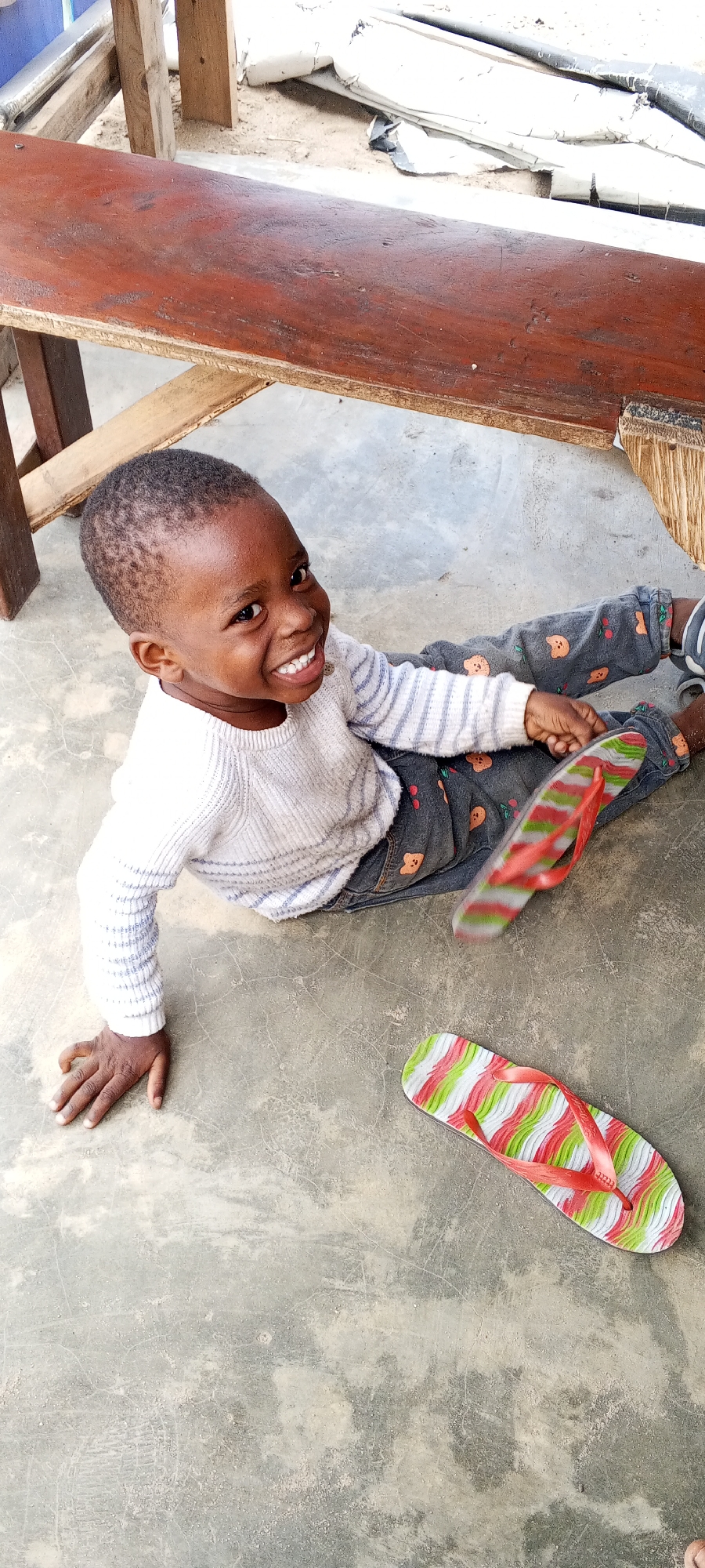 A young child with dark skin and short dark hair is captured sitting on a concrete floor in Lagos, Nigeria. The child looks directly at the camera with a wide, joyful smile, displaying their teeth. They are dressed in a light-colored, possibly white or light grey, long-sleeved sweater with subtle horizontal stripes, paired with dark grey pants adorned with light-colored cherry and bear patterns.
One colorful flip-flop, featuring wavy stripes of red, green, and white with a red strap, rests on the concrete floor to the child's right. Another identical flip-flop is blurred in motion near the child's feet, suggesting it is being played with or has just moved. The child's left hand is placed flat on the concrete.
In the upper background, a rustic wooden bench or table with a reddish-brown top and visible wooden supports is partially visible. Behind the bench, light-colored, possibly crumpled fabric or plastic materials are discernible on the ground. The scene is well-lit by natural light, indicating it is daytime. The child's expression conveys clear happiness and playfulness. No text is visible in the image.