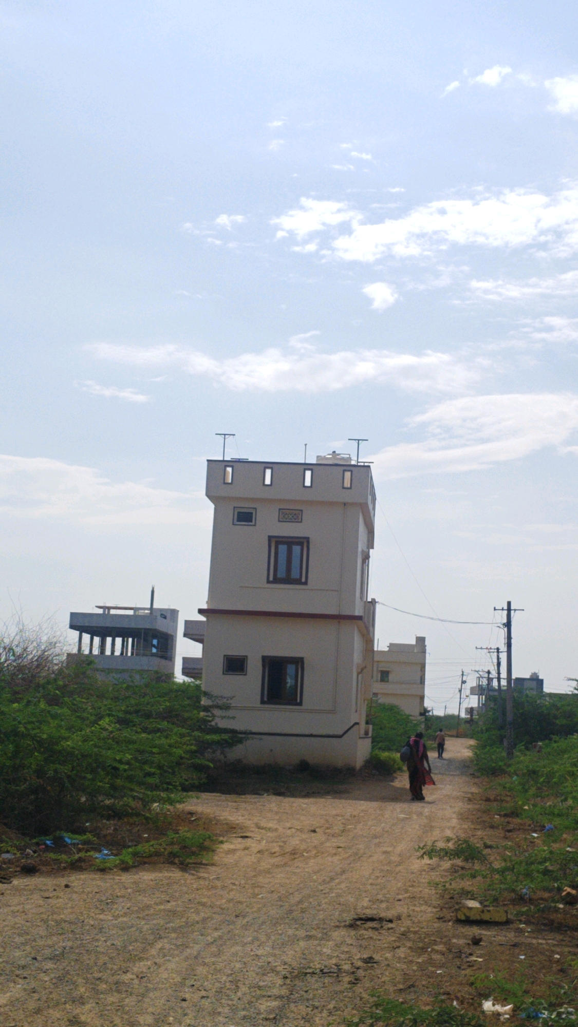 The image depicts a dirt road in Pamur, India, with several buildings visible in the background. The primary focus is a tall, narrow, cream-colored building resembling a tower with a flat roof that has some architectural elements on top. To the left of this tower is another building, partially constructed, with a visible concrete structure and support pillars. Further down the road, other buildings and utility poles are present. The environment is characterized by dry, dusty ground, sparse vegetation, and a light blue sky with scattered clouds. Two individuals are walking away from the camera on the dirt road, one woman in a colorful sari carrying a bag over her shoulder and a second, more distant figure. The scene appears to be in a developing or rural area.