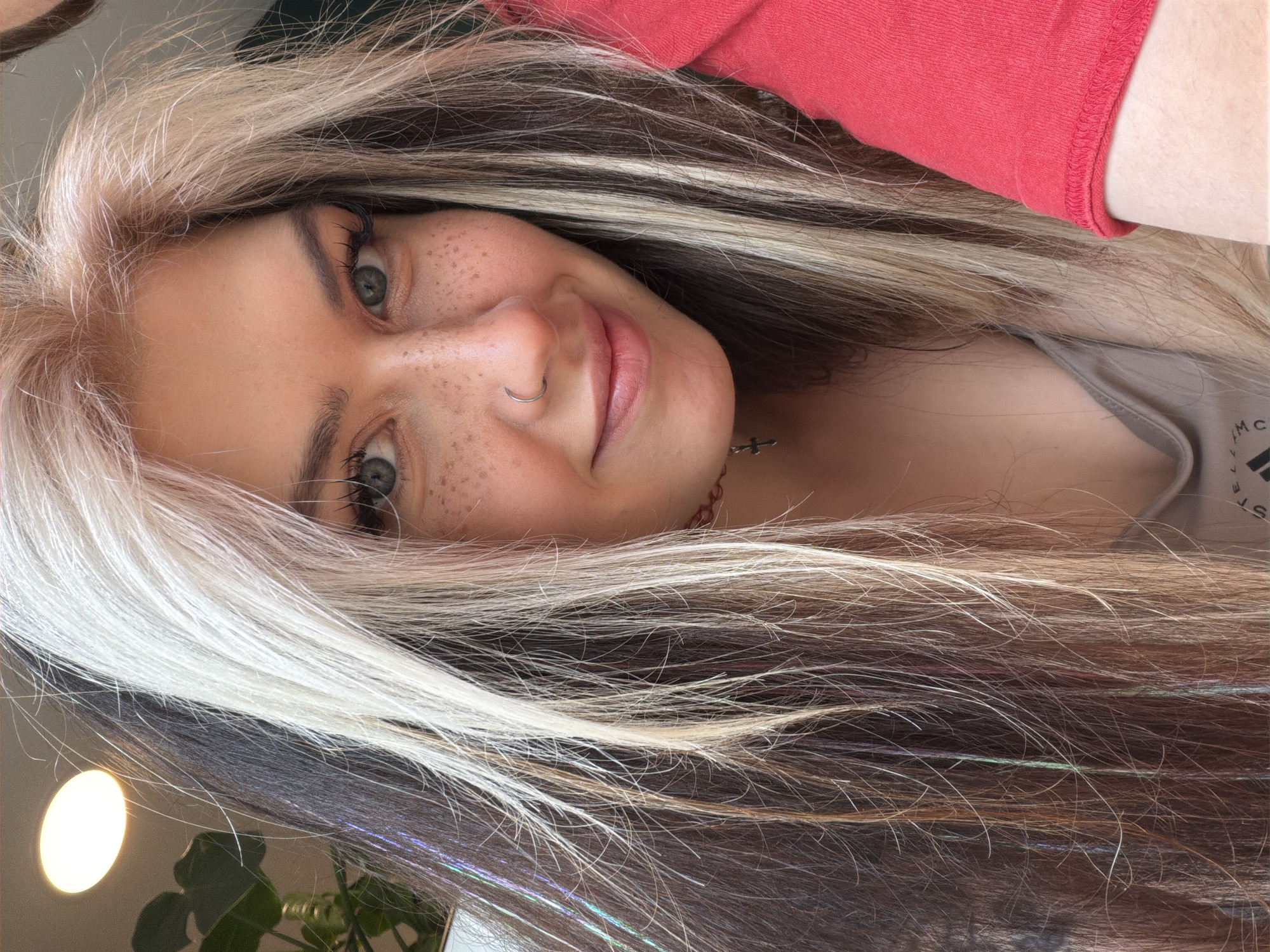 A close-up, angled portrait of a young woman with fair skin, freckles, and a nose ring. Her eyes are light-colored. She is smiling slightly. Her hair is long and multi-toned, with blonde and darker brown shades. Some strands appear to have iridescent tinsel woven in. She is wearing a red shirt in the upper portion of the frame and a tan shirt underneath. A small cross pendant is visible on a necklace. Part of a plant with green leaves is visible in the lower left corner. The location is not identifiable.